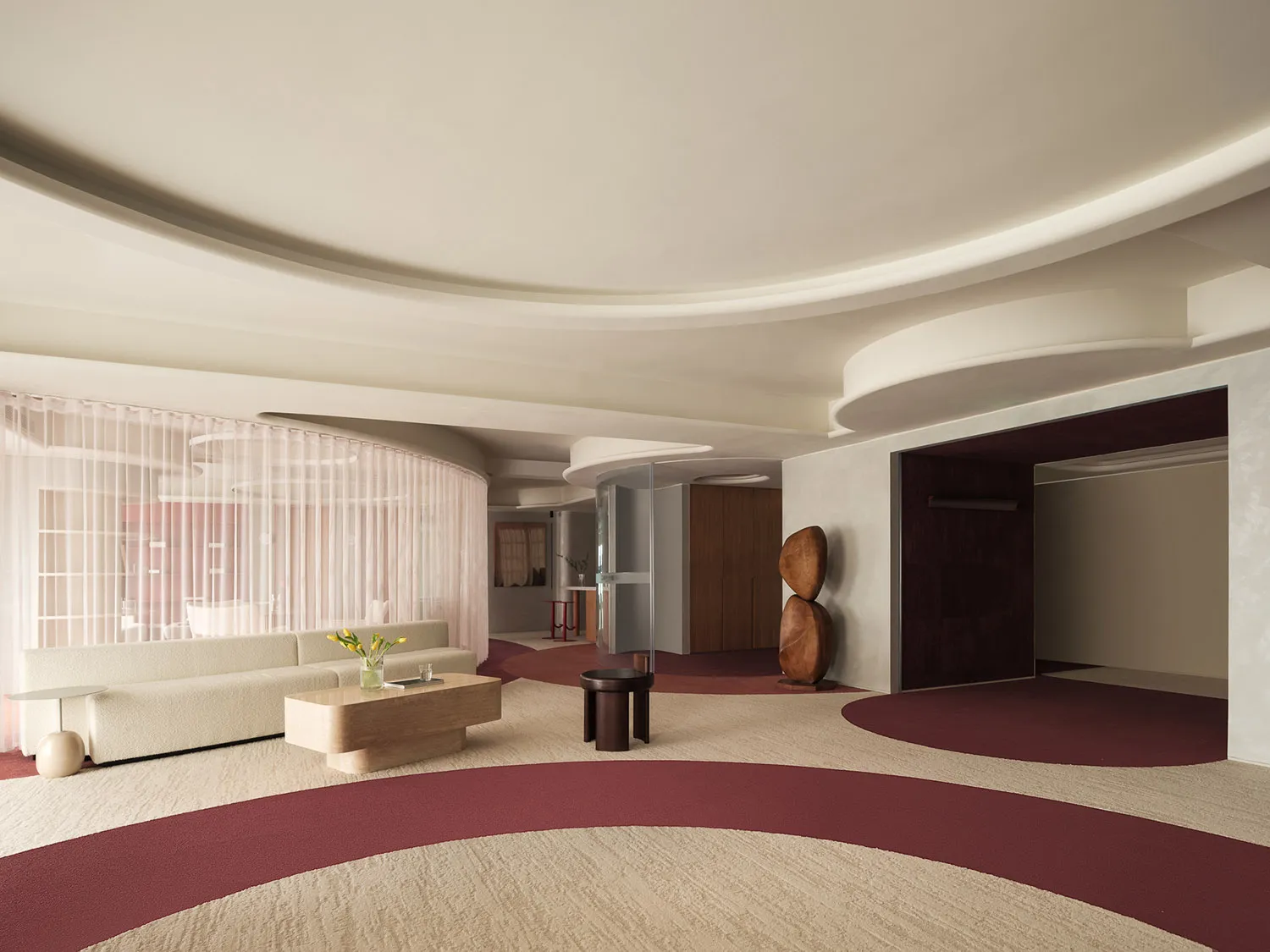 A modern lobby with white sofas, a wooden coffee table, and a textured carpet in shades of red and beige. The ceiling feature