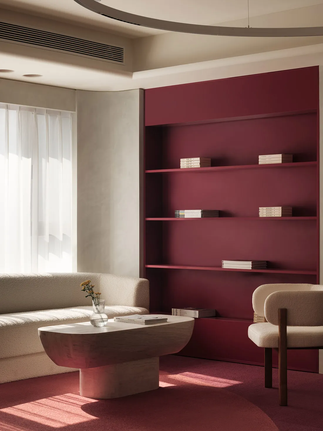 A minimalist living room with a red shelving unit and white furniture, featuring a table, a chair, and a window with sheer cu