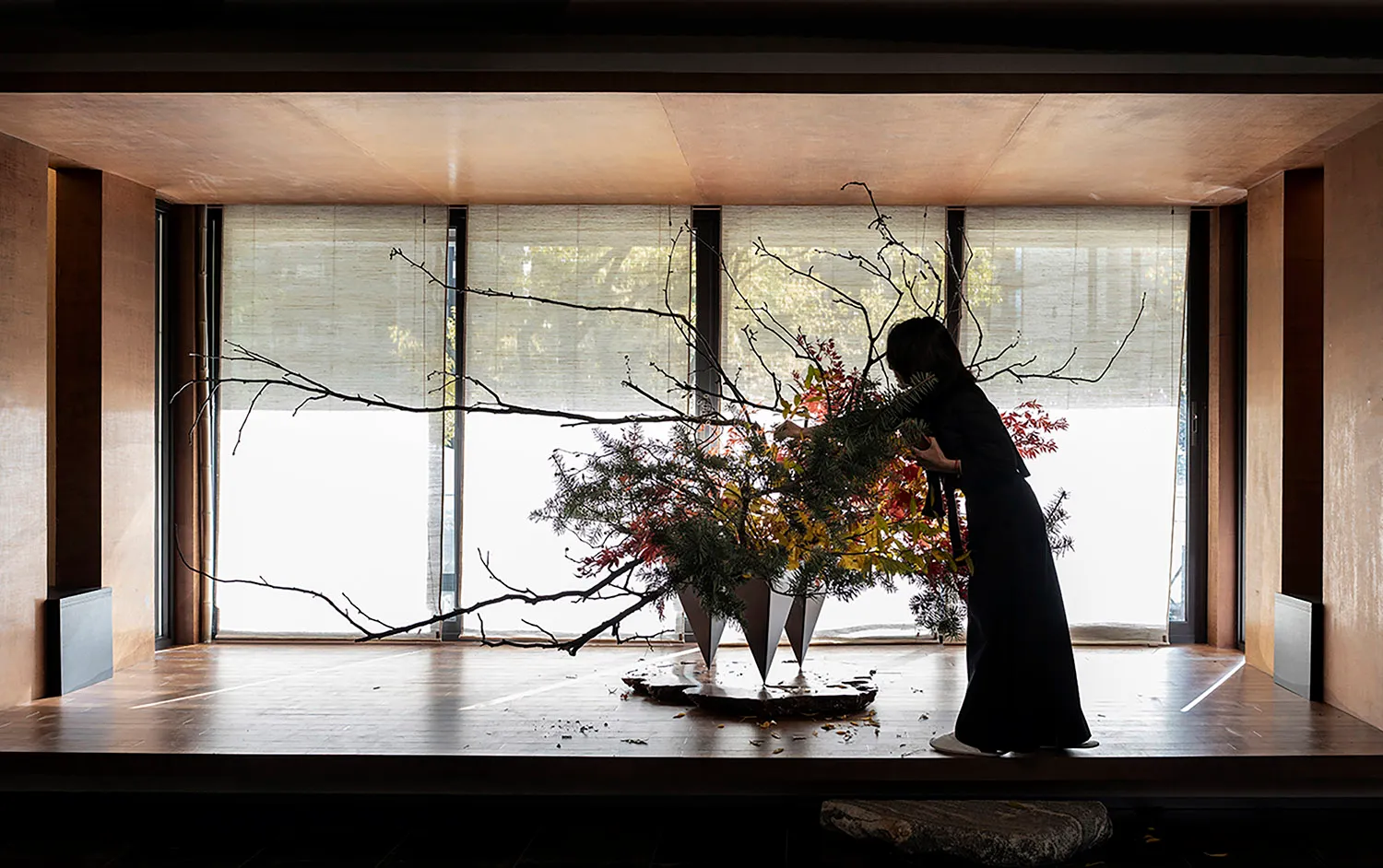 A woman in a black dress arranging a floral arrangement with wooden branches and foliage in a concrete space
