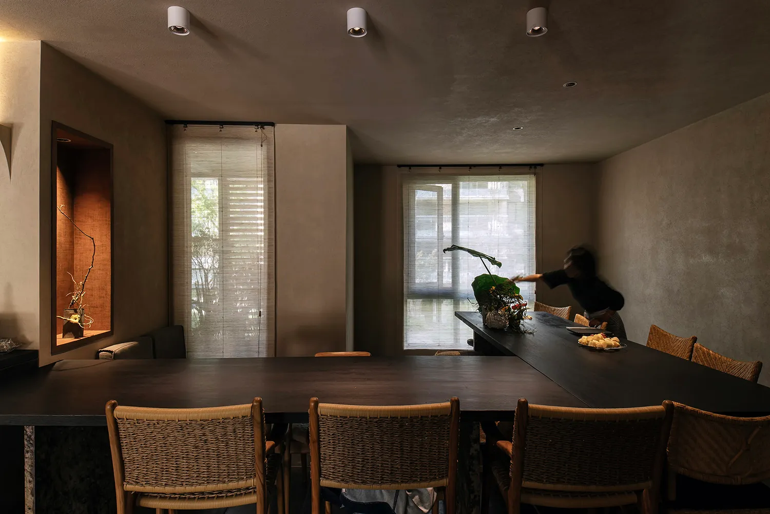 A modern dining room featuring a black rectangular table surrounded by wooden wicker chairs, with a person adjusting the blin