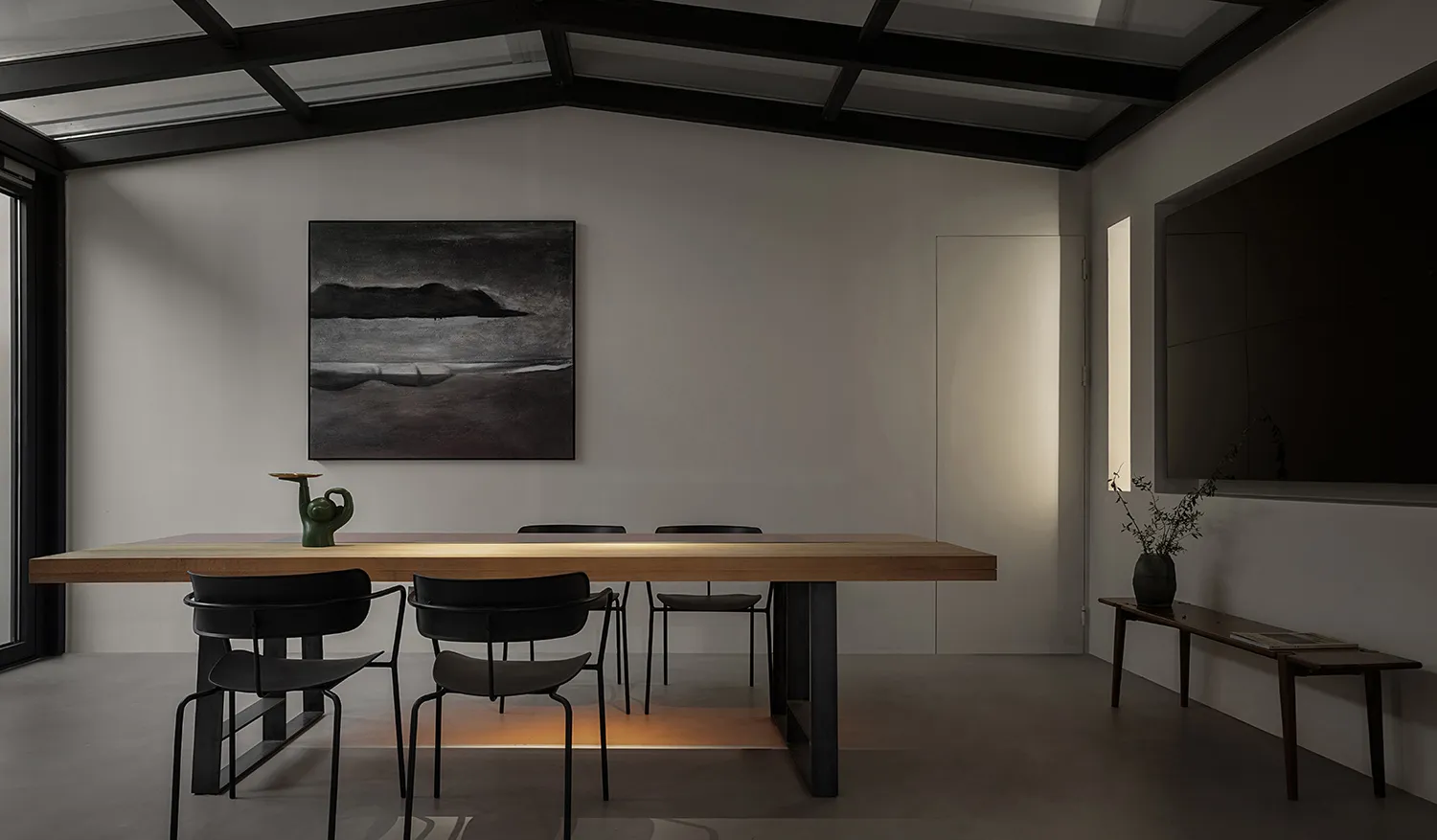 A minimalist living room featuring a wooden dining table with black chairs, a black and white abstract painting, and a small