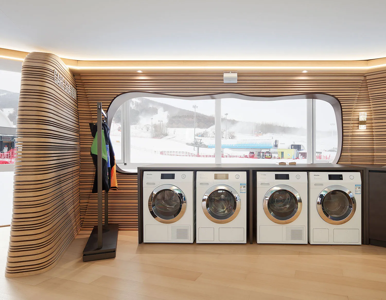 A modern laundry room featuring wooden walls with a row of washing machines next to a window looking out to a snowy landscape