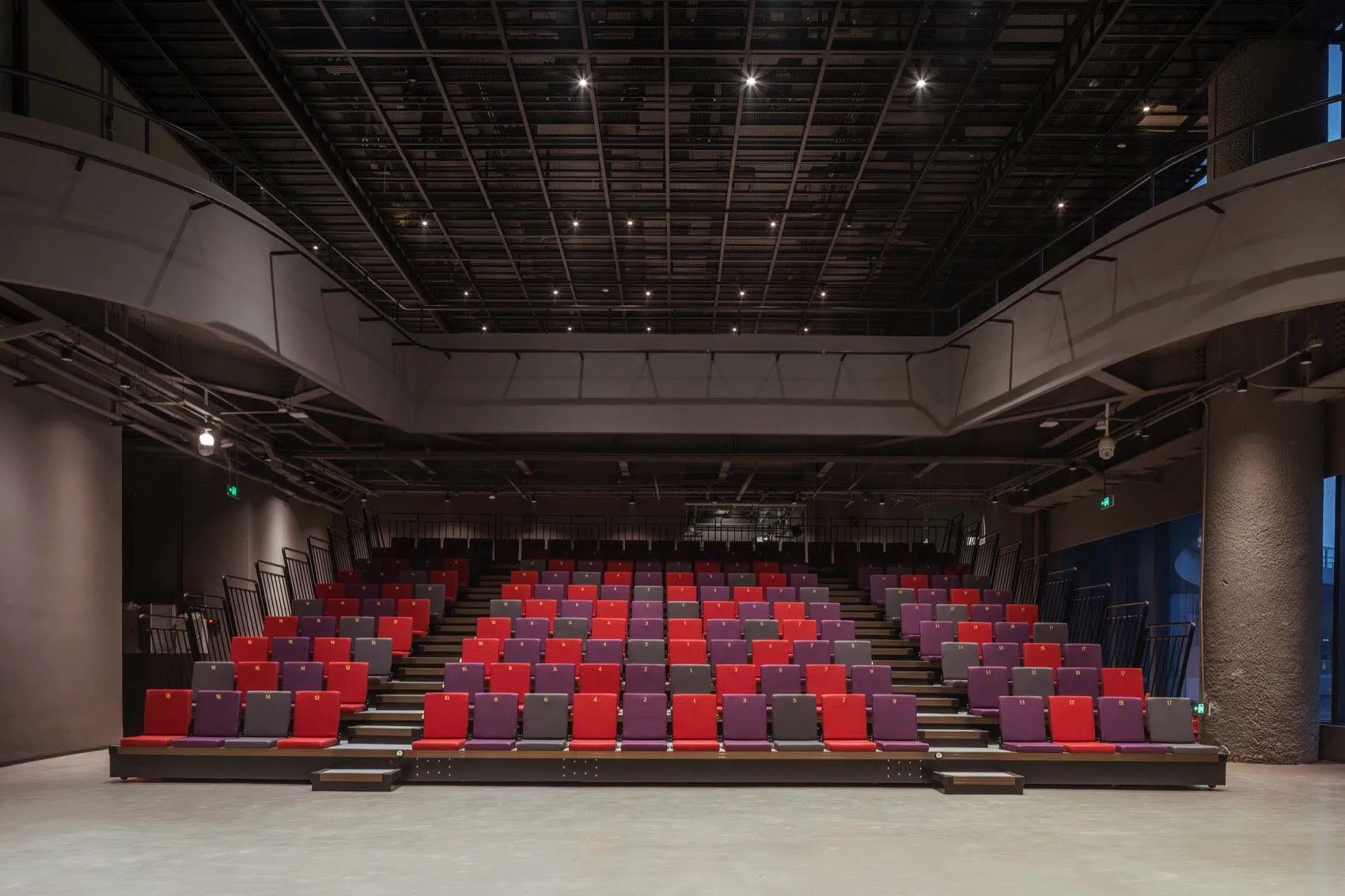 Empty industrial-style auditorium with metal seats, black ceiling structure, and concrete floor.