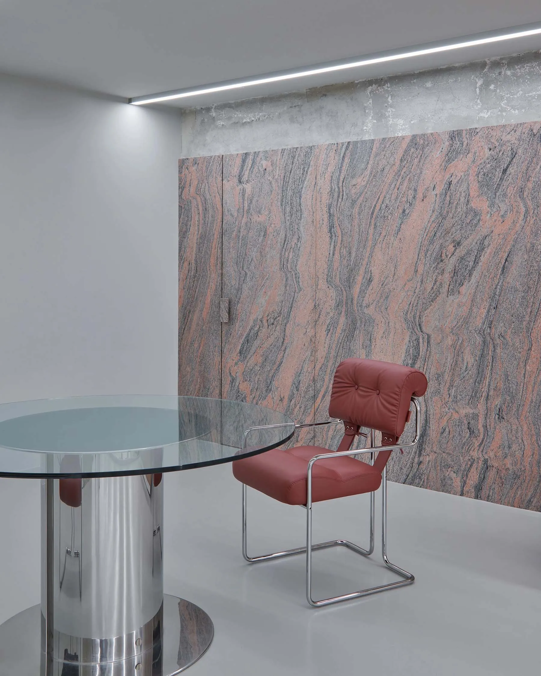 A modern office space with a sleek, curved glass table, a red leather chair with metal frame, and a concrete wall with marble