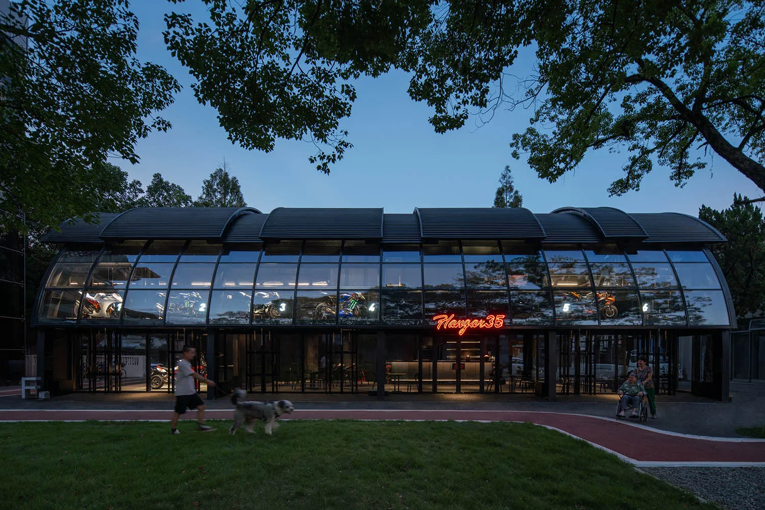 A modern restaurant exterior with large glass windows, illuminated signage, and a dog walking on the grass.