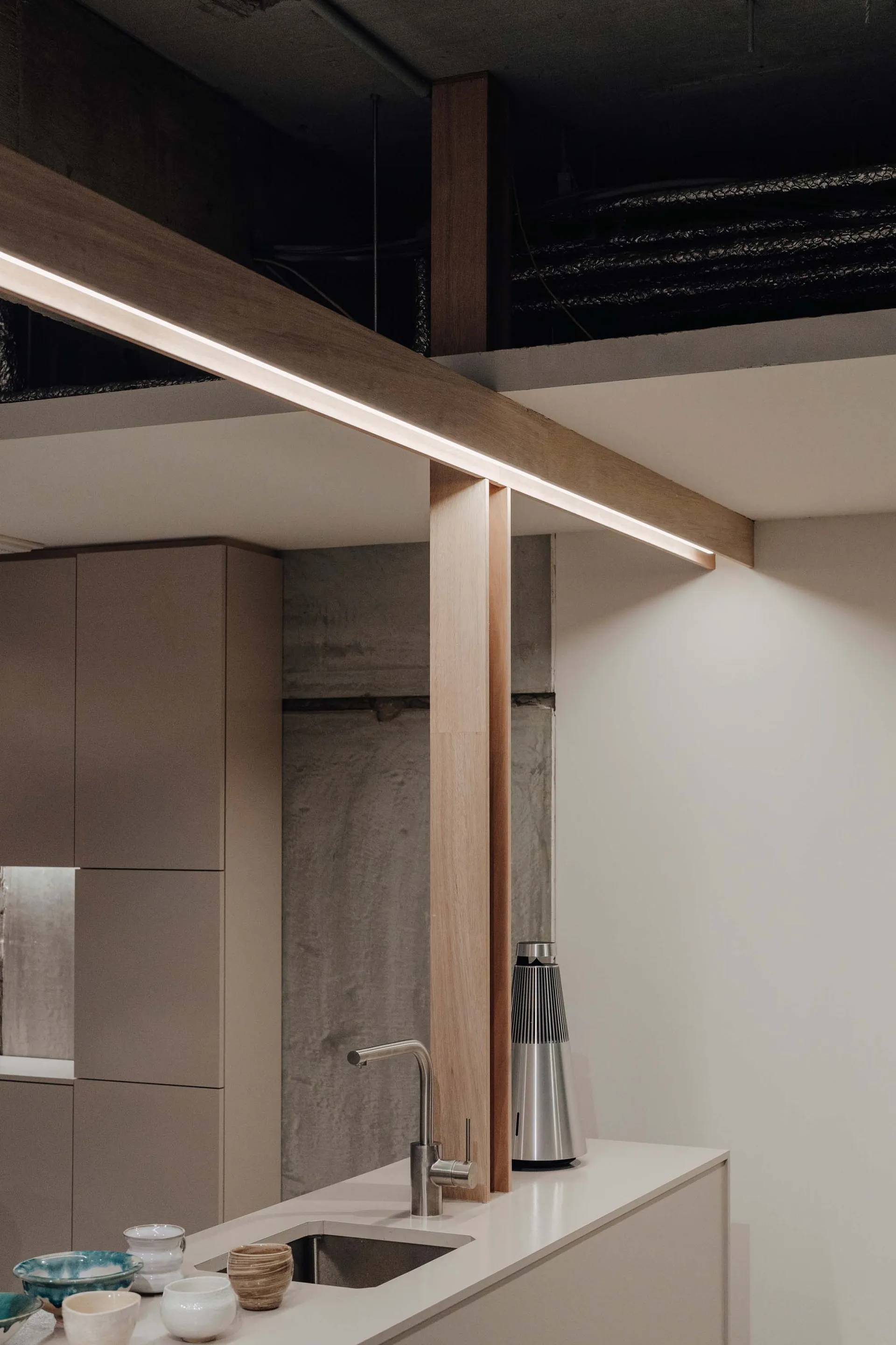A minimalist kitchen with concrete walls and white countertops features a wooden beam with integrated lighting and a sleek st