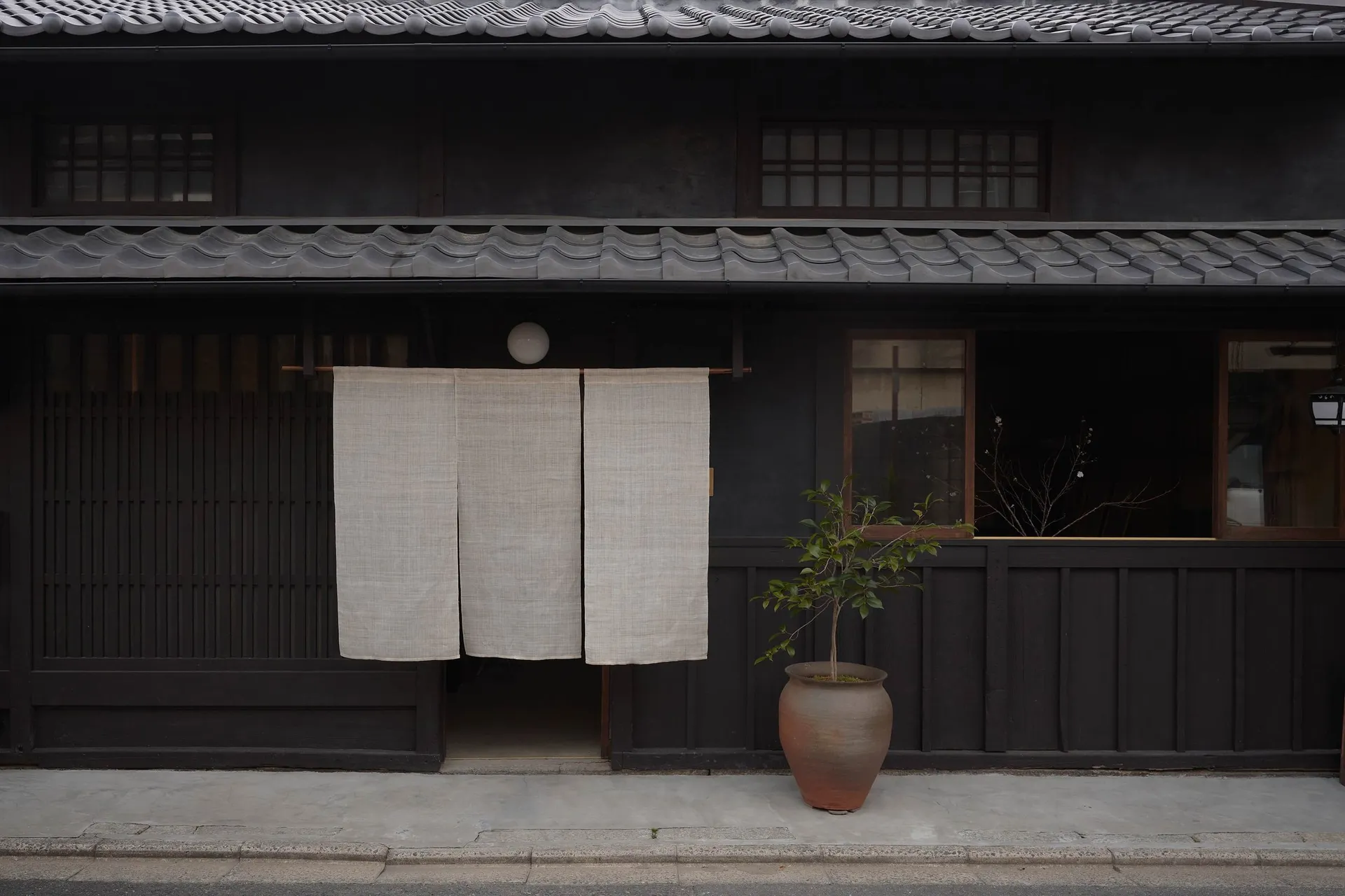 A traditional Japanese storefront with wooden shutters, concrete steps, and a large fabric curtain. A small plant in a pot ad