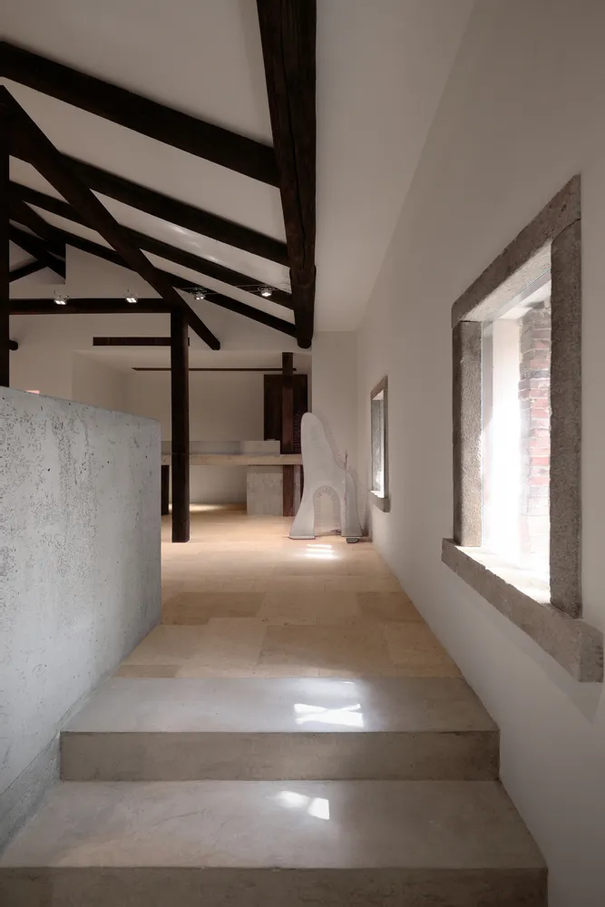 A modern interior space with concrete walls and flooring, featuring exposed wooden beams and a large window allowing natural