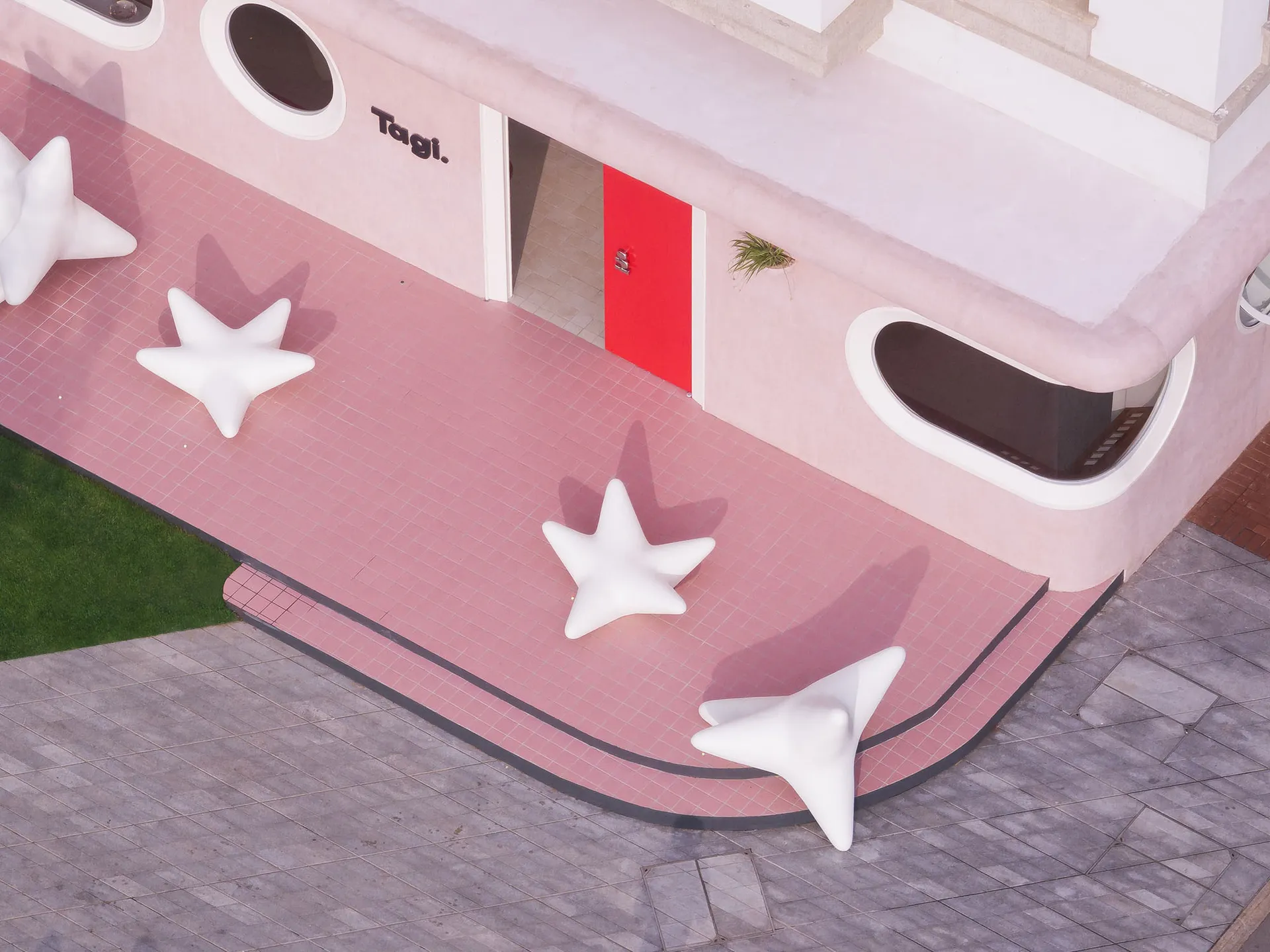 A modern retail exterior with a pink building facade, white modern sculptures, a red door, and green plants.