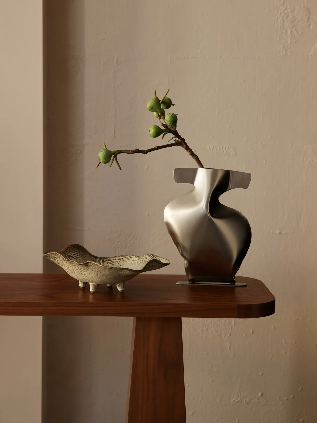 Two decorative vases on a wooden table, one metallic with abstract design and one ceramic with ruffled edge.