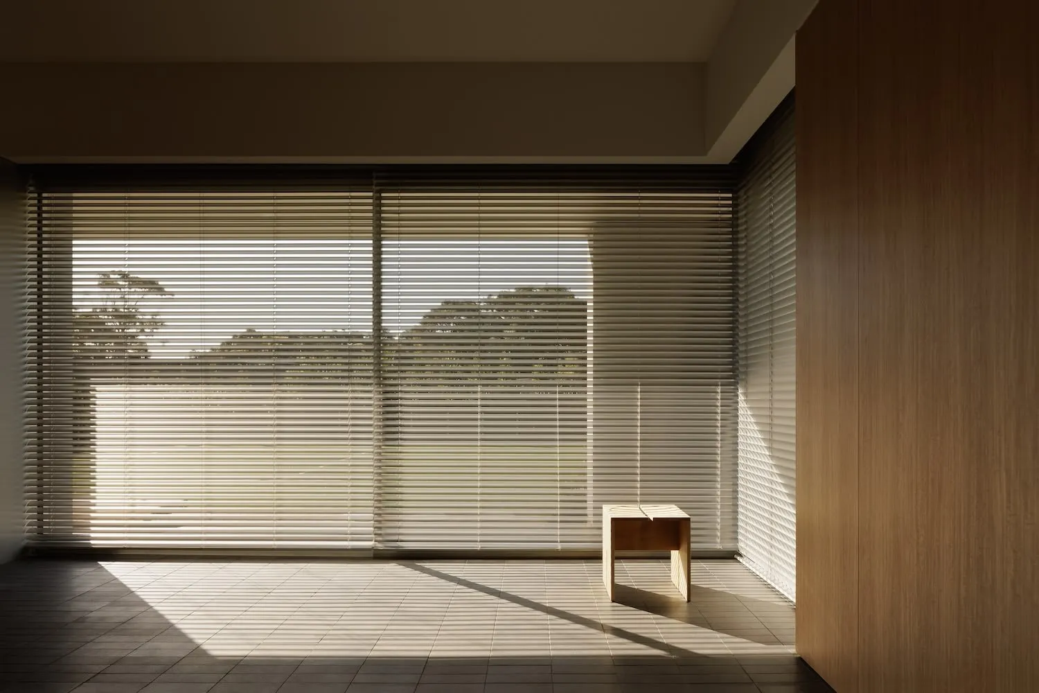 Minimalist interior room with wooden floors and a single wooden bench in front of a large window with blinds.