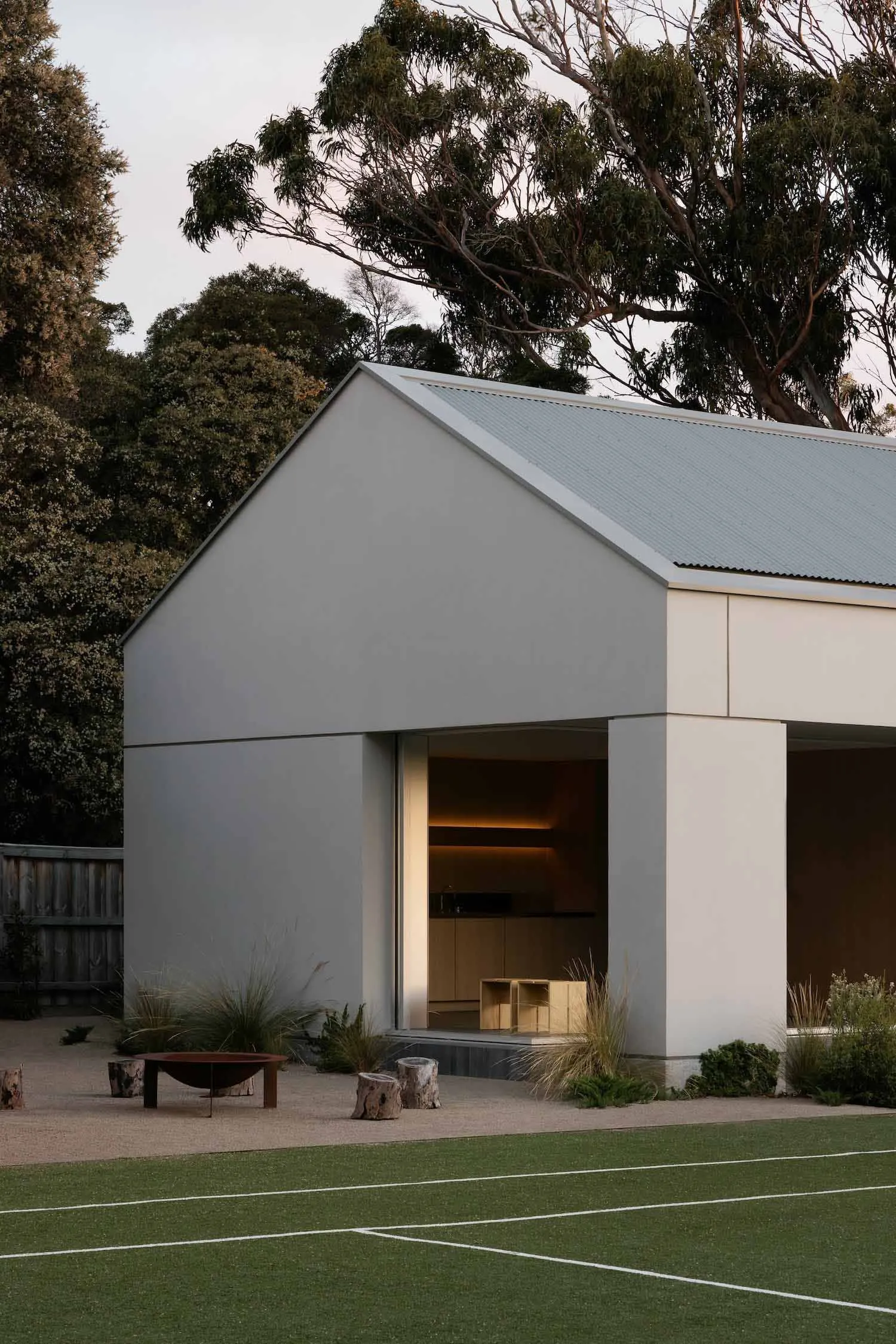 A modern residential courtyard with a concrete and steel-cladding exterior, featuring a fire pit and natural foliage.