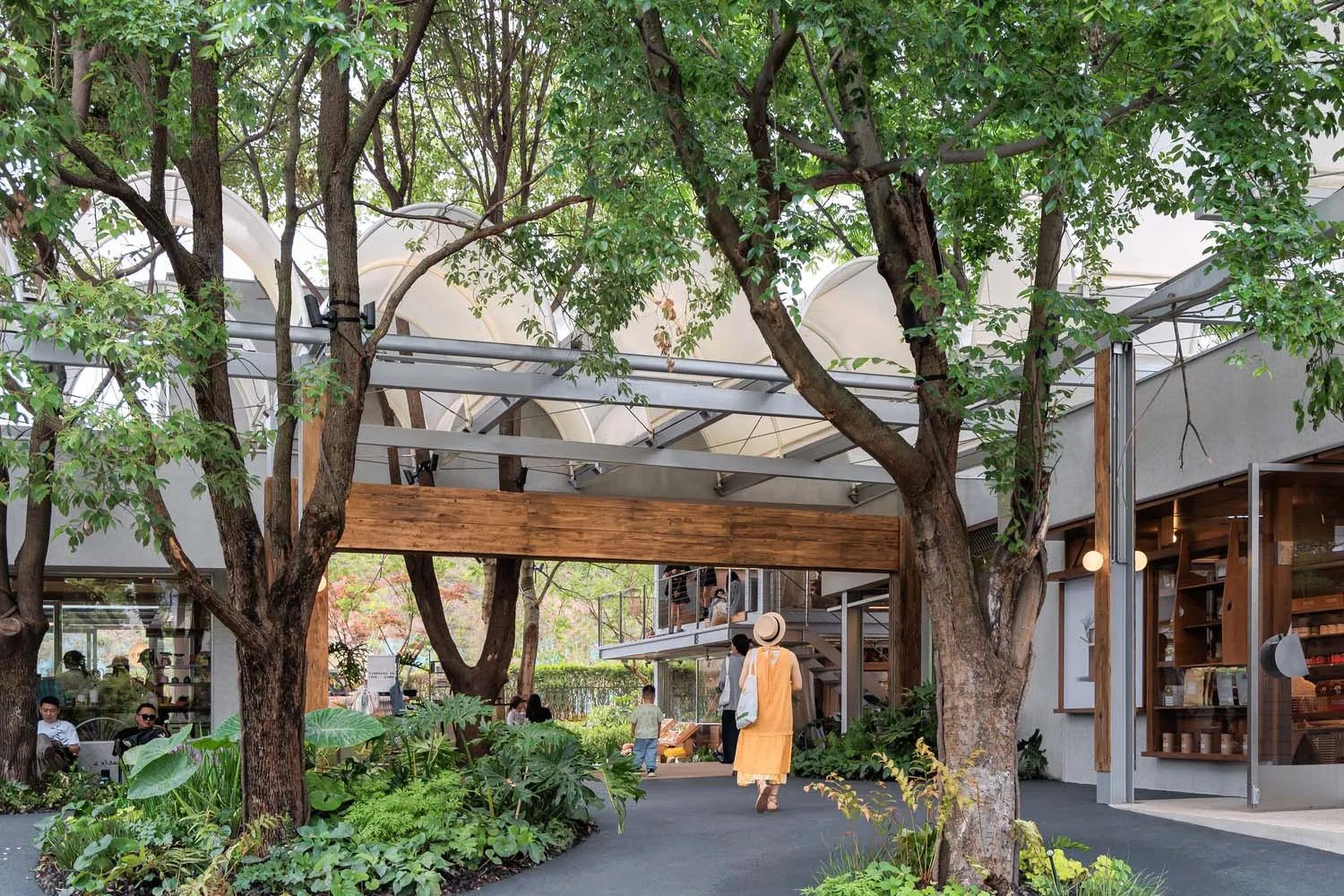 Outdoor shopping center with wooden walkways, glass walls, and concrete structures surrounded by greenery.
