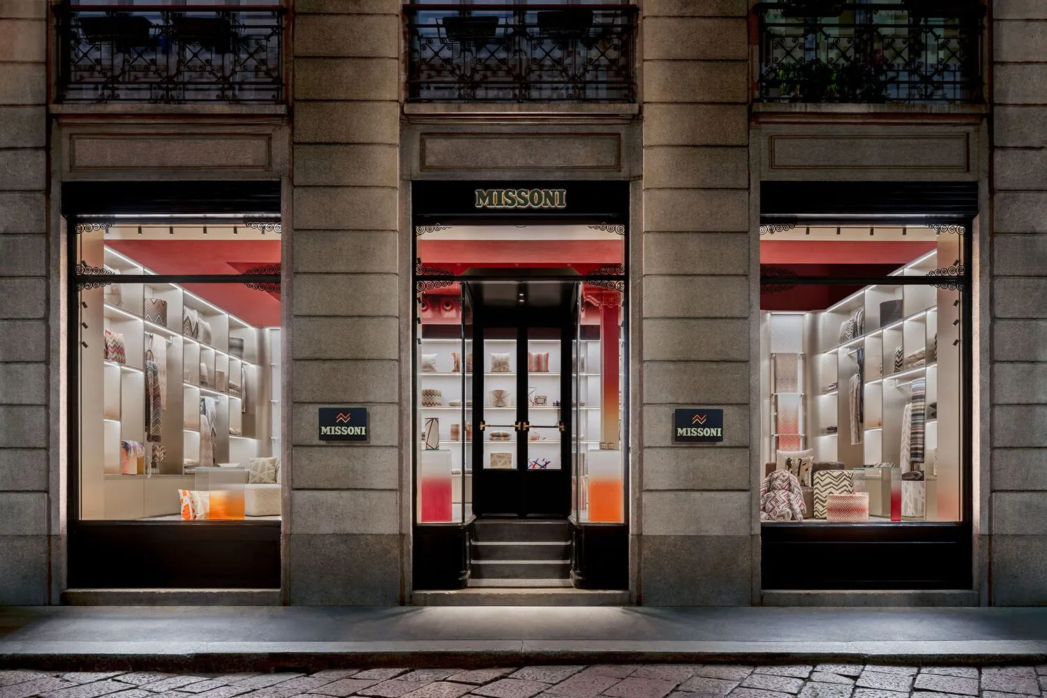 A modern storefront with glass windows displaying retail items, featuring a metal door and exterior wrought iron railing.