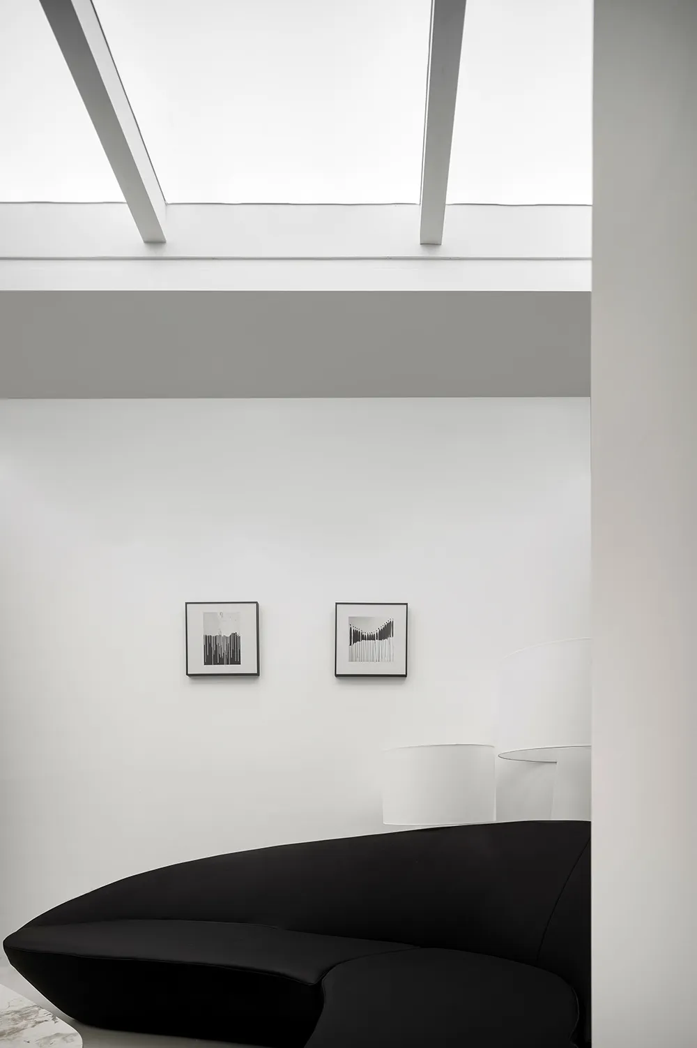 A minimalist living room featuring a sleek black couch on a white floor, with a skylight and framed pictures on the wall.