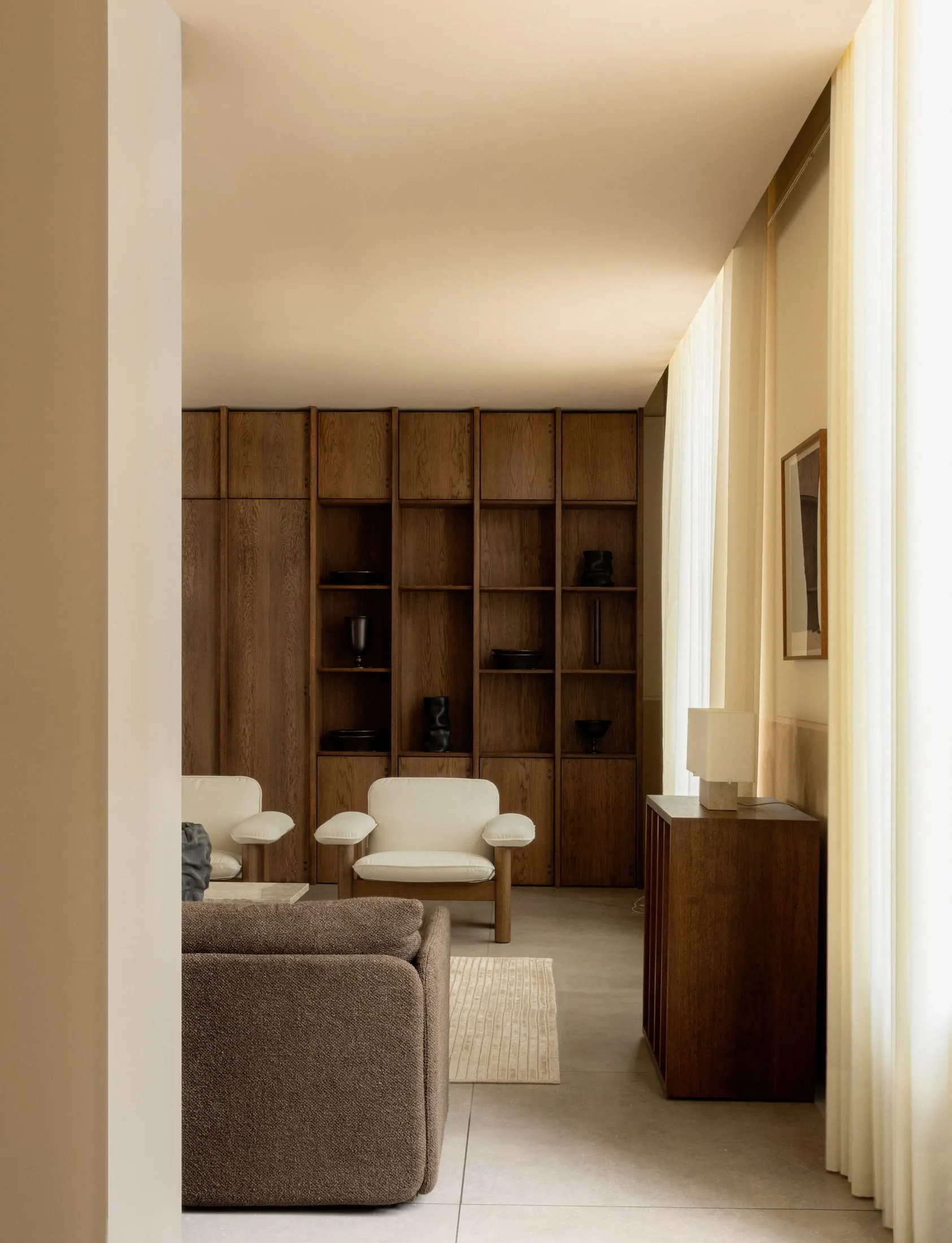 A minimalist living room with neutral tones, featuring a brown sofa, white chairs, and a wooden bookshelf.
