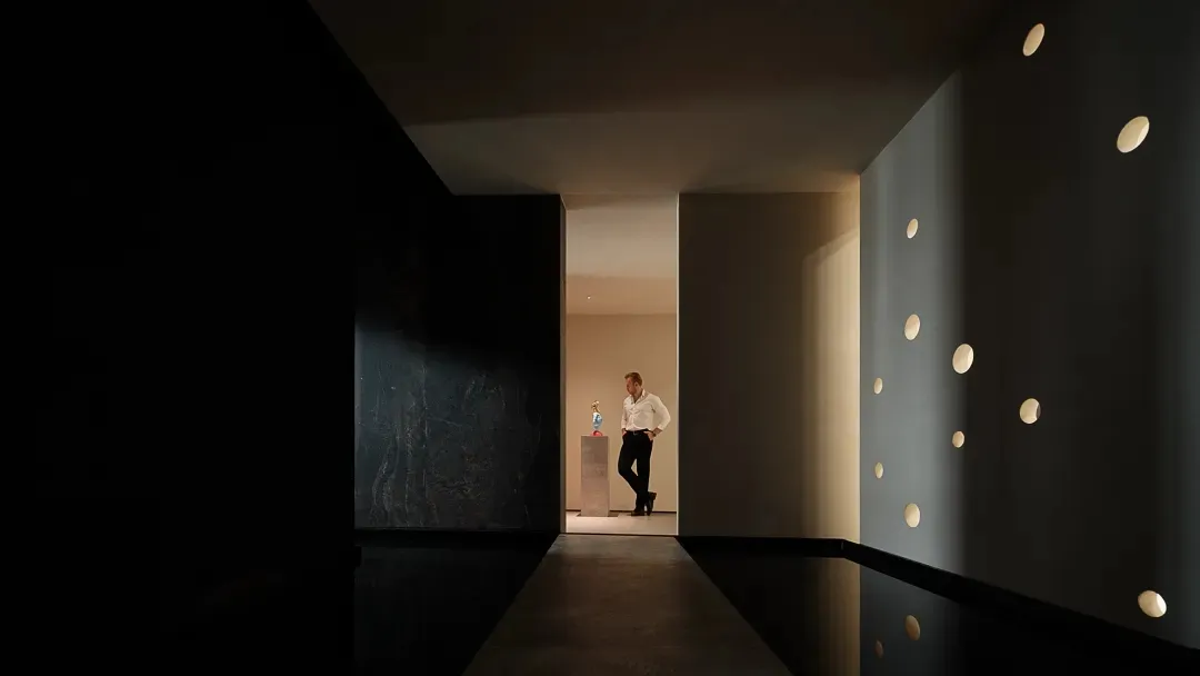 A modern, minimalist corridor with concrete walls and floor, featuring a series of circular light fixtures on the right wall.