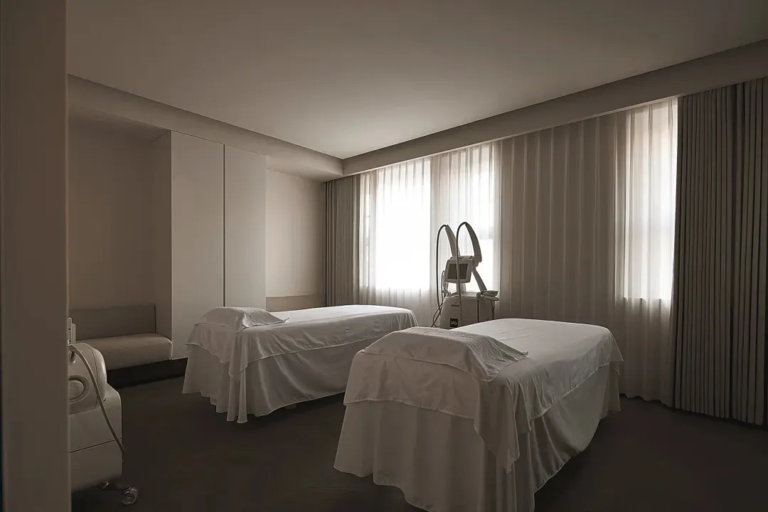 A minimalist spa room featuring two beds draped in white linen, with large windows and soft lighting.