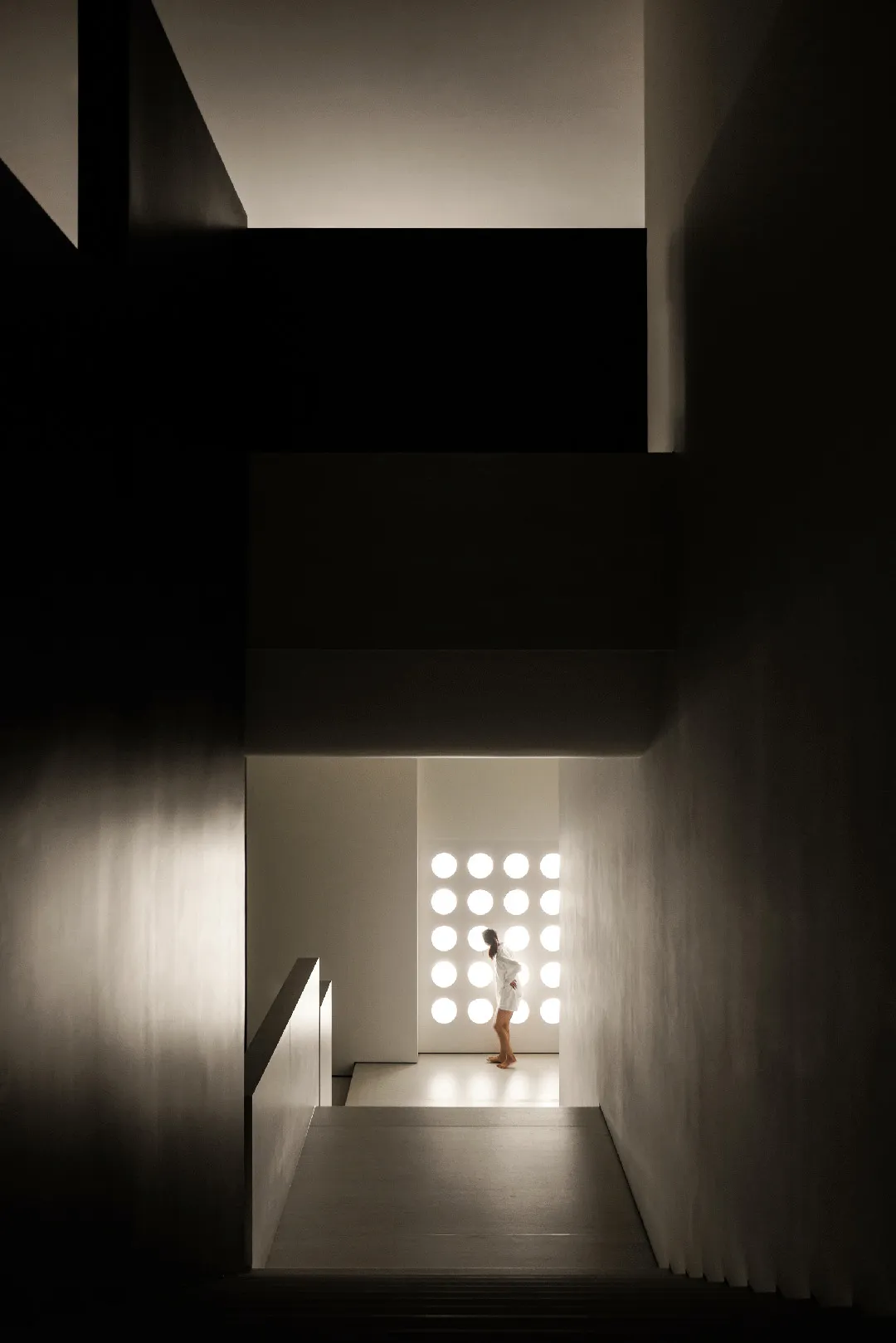 A minimalist interior space with a concrete wall and a person standing in front of a lighted wall featuring circular patterns