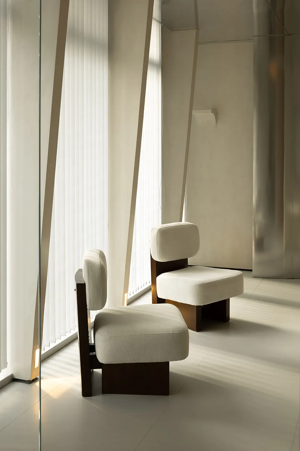A minimalist living room with white cushions on wooden chairs and stools, featuring vertical blinds and natural light.