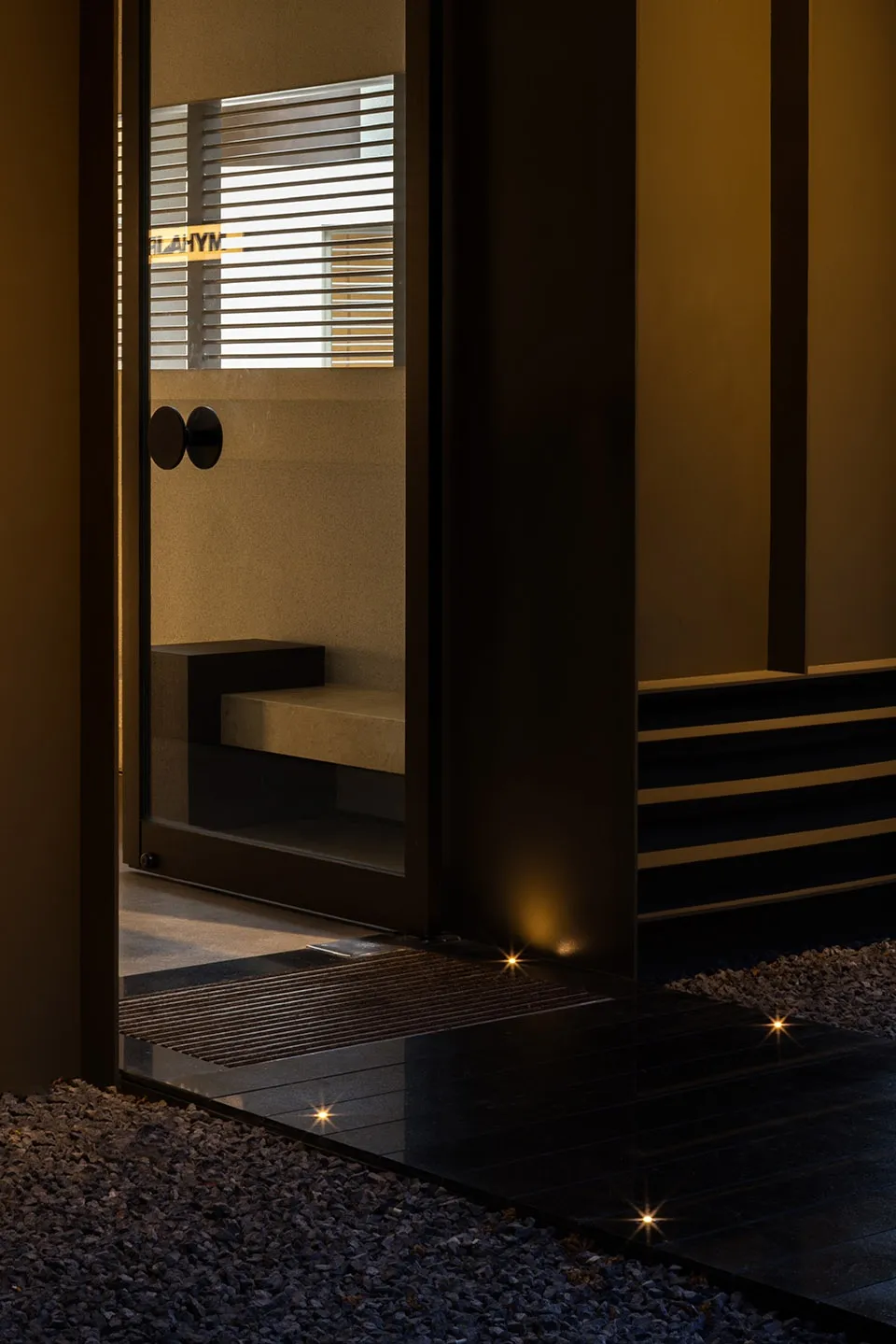 A modern entrance featuring a glass door, concrete steps, and gravel pathway illuminated by warm lights.
