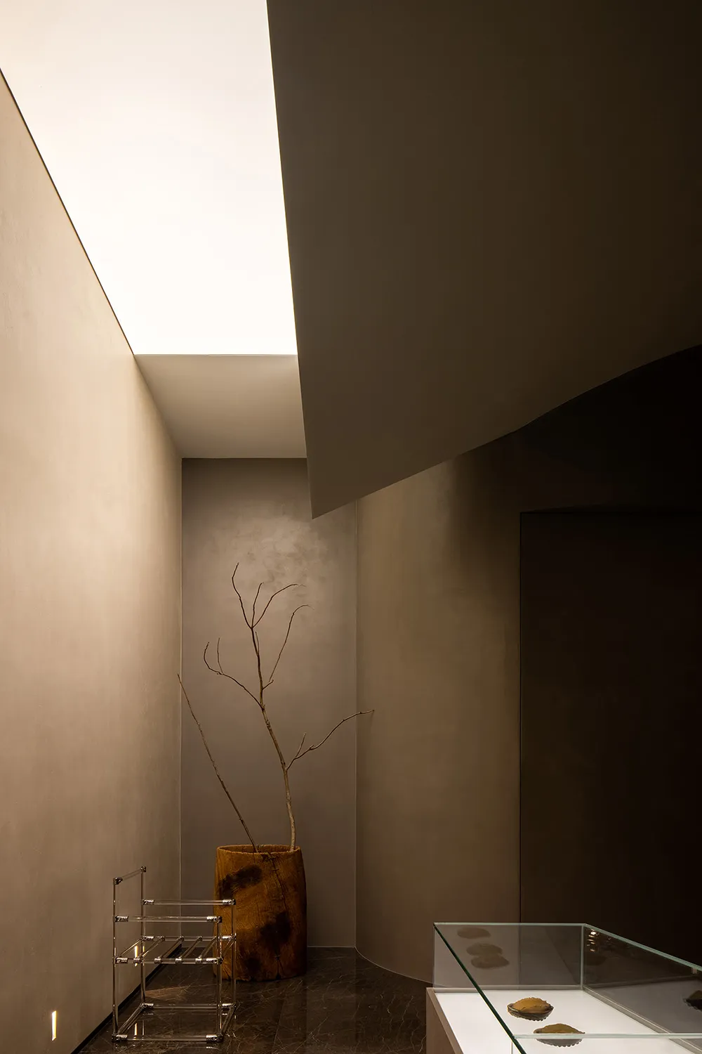 A minimalist hallway with modern concrete walls, a glass display case, and a single wooden vase with branches.