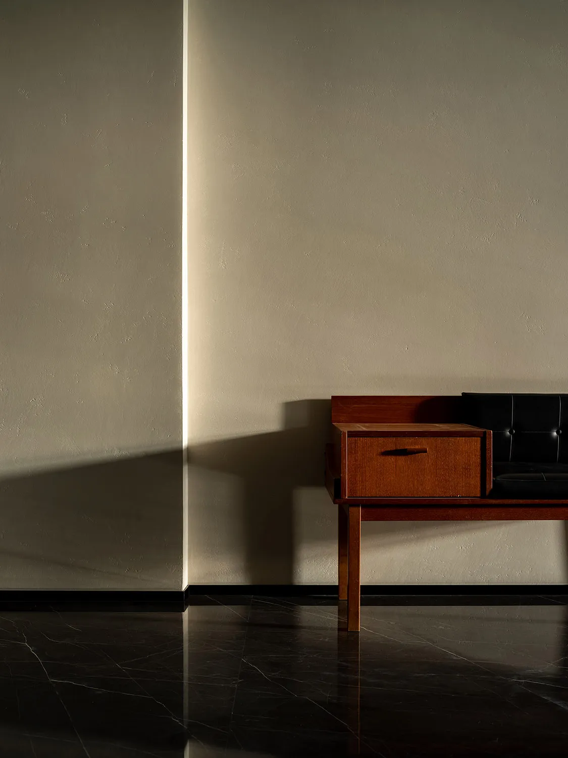 A minimalist interior with a wooden table and black leather seating against a plain wall and marble floor.