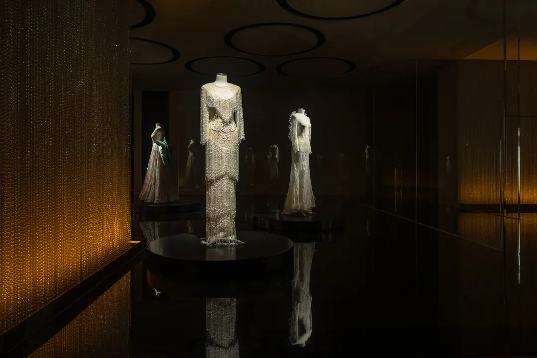 An interior image showcasing a glass exhibit space with illuminated mannequins and dresses.
