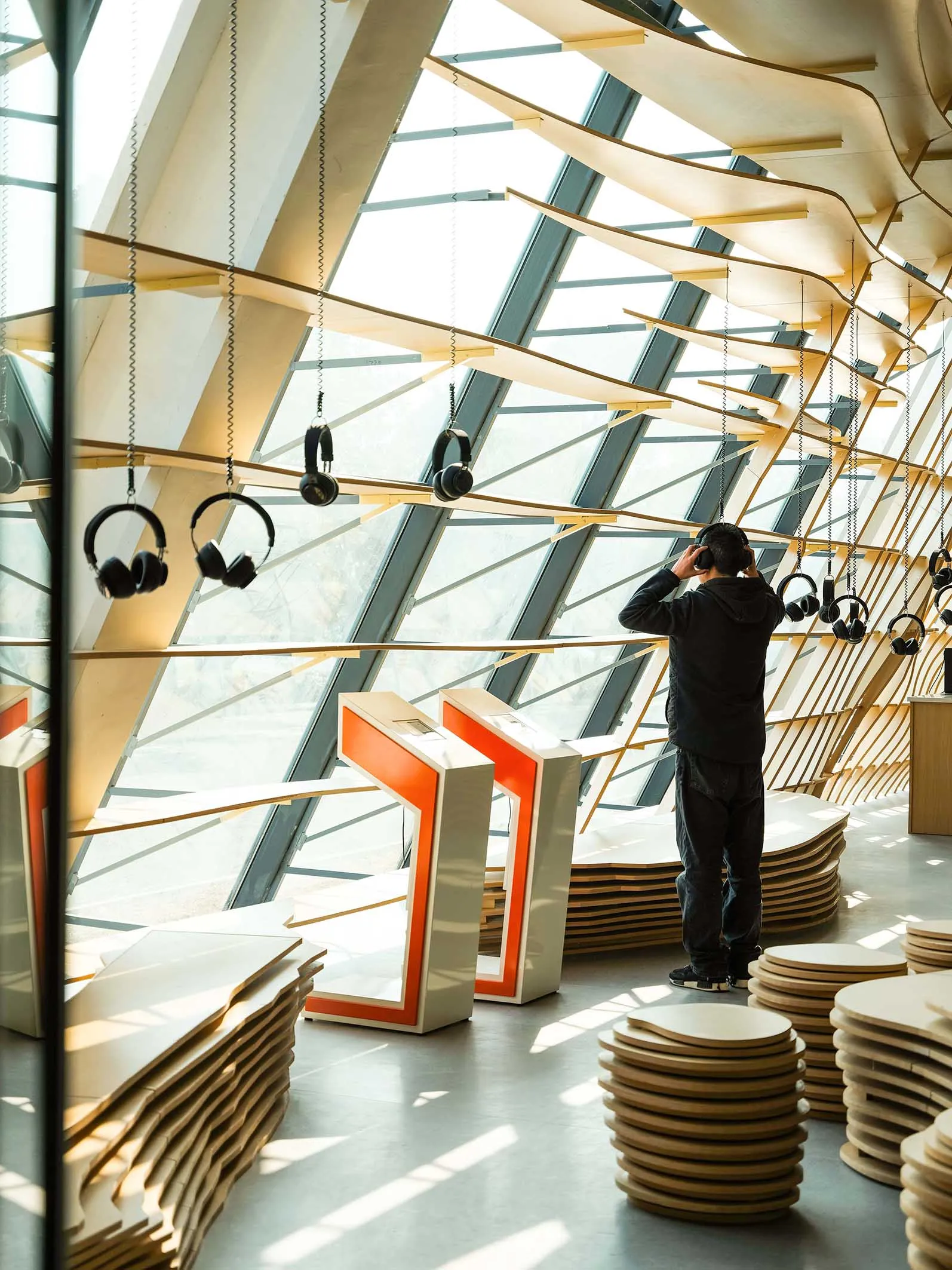 A person wearing headphones in a spacious, industrial exhibition hall with wooden structures and hanging headphones.