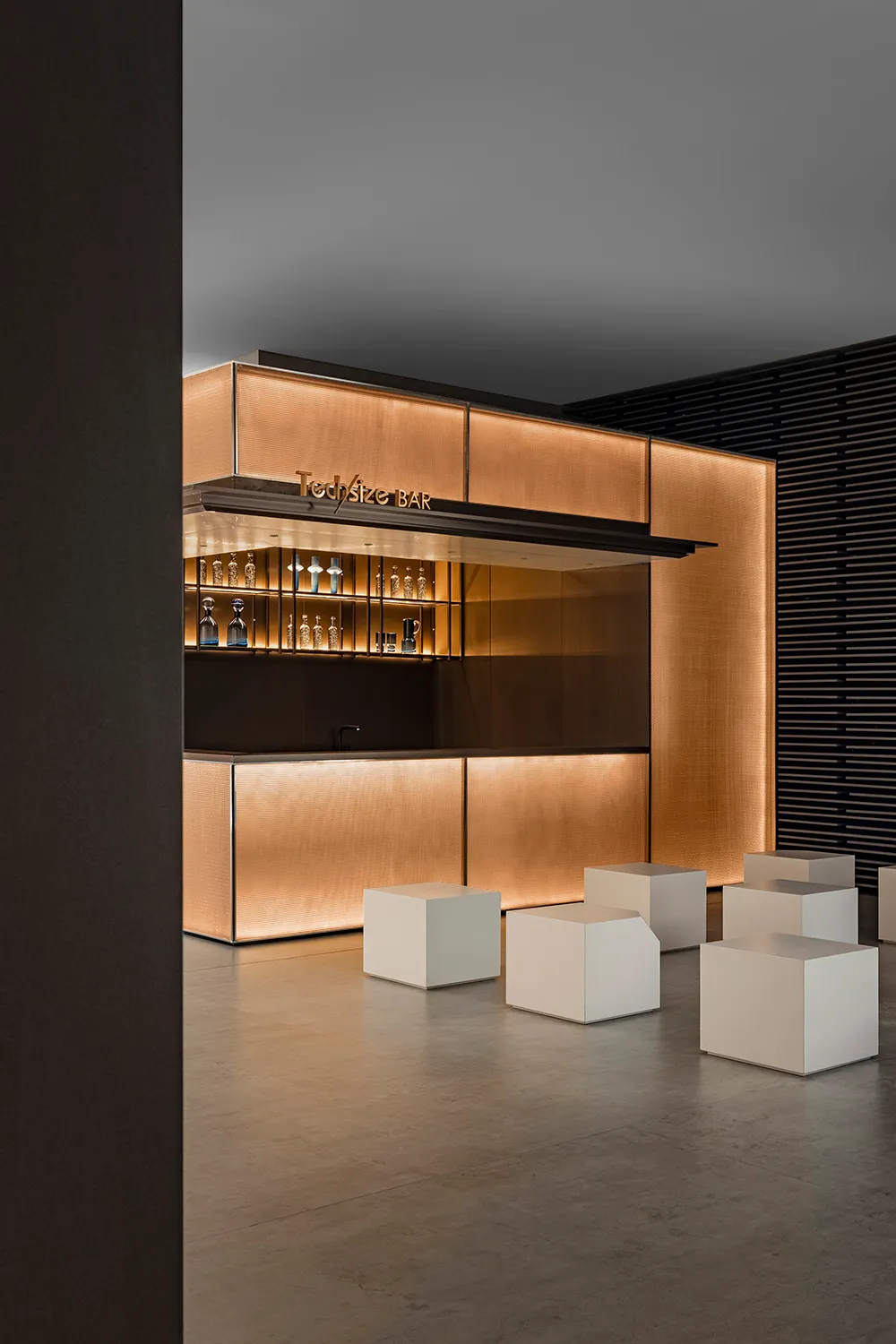 Modern bar counter featuring metallic texture, illuminated with warm lighting, and accompanied by minimalist seating.