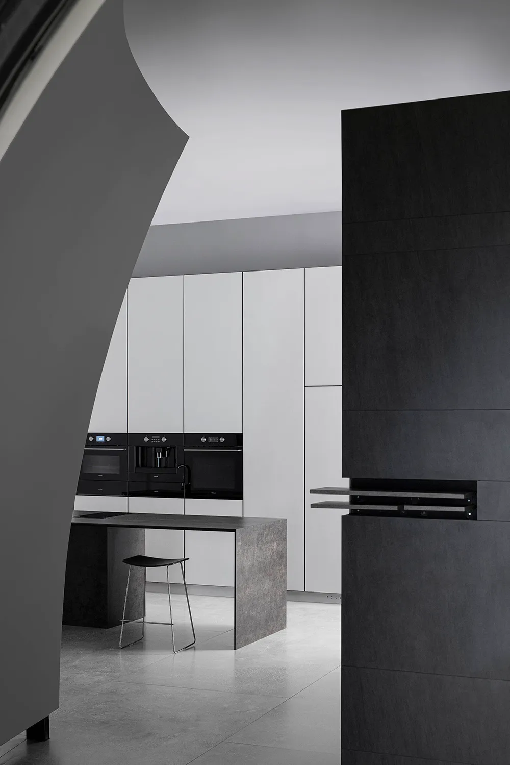A modern, minimalist kitchen with white cabinets, concrete countertops, black appliances, and a black and metal bar stool.