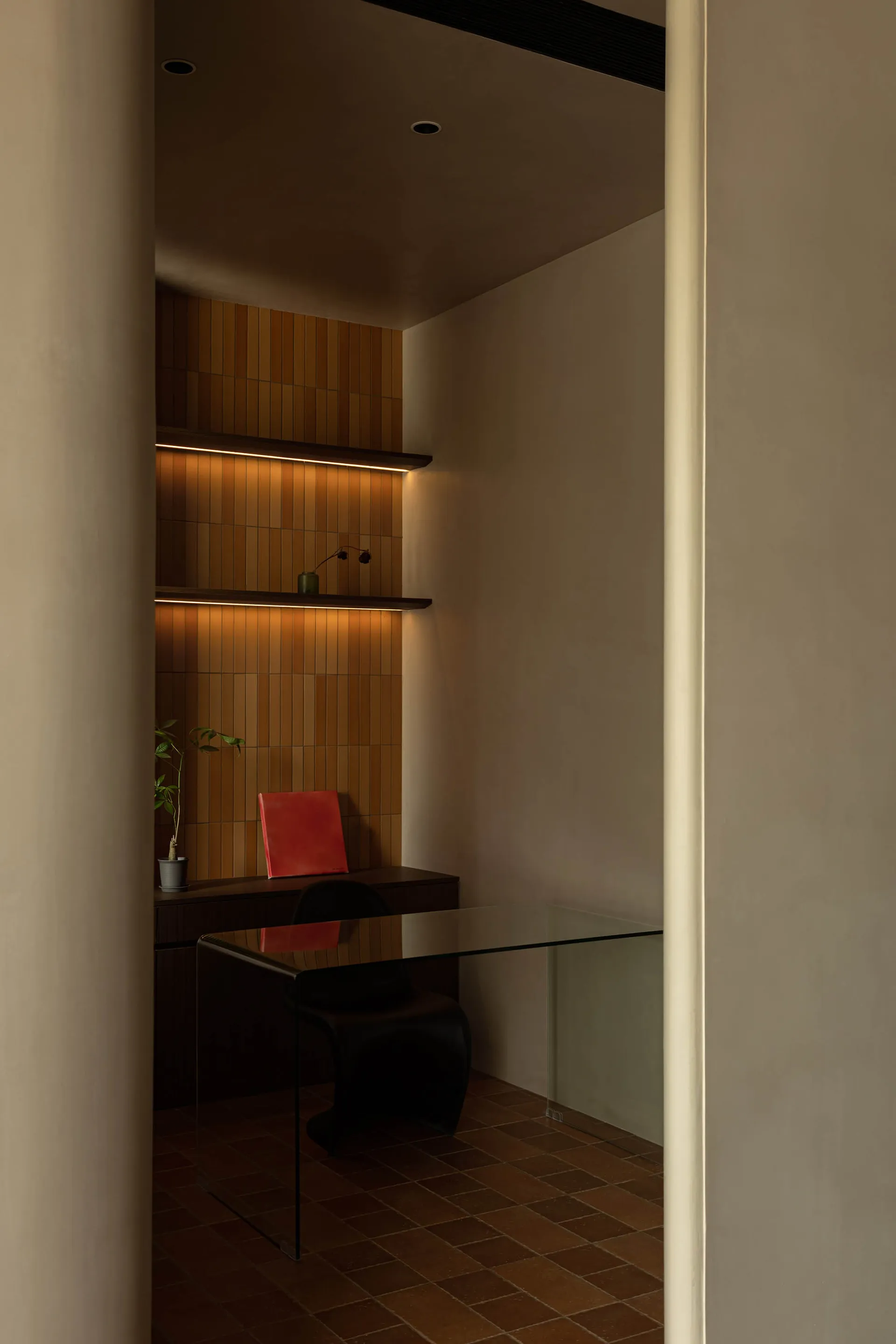 A modern minimalist office with glass desk, red book, black chair, plant, and shelving with books and decorative items.
