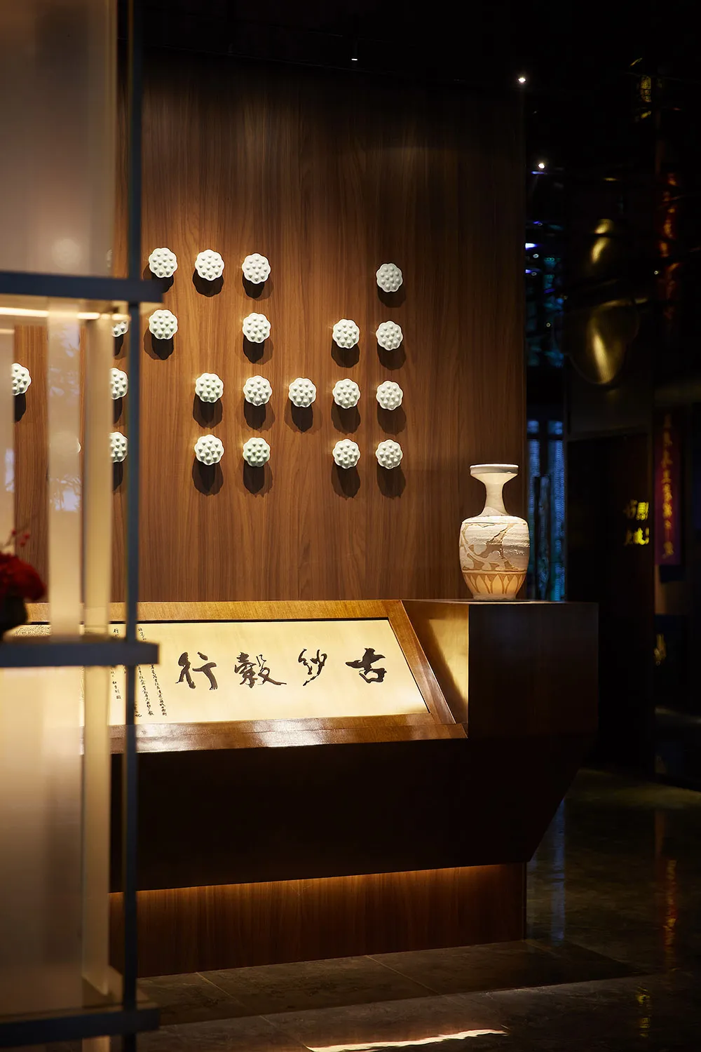 A wooden wall adorned with white ceramic installations, featuring a vase and calligraphy on a wooden stand.