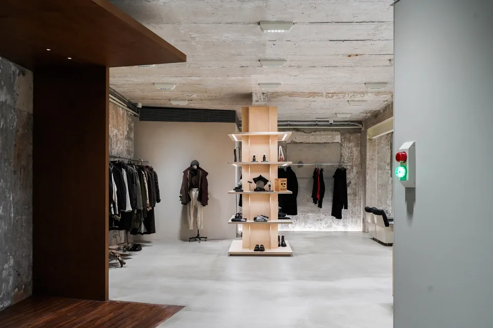 A minimalist clothing boutique with concrete walls and floors, featuring wooden shelves and mannequins displaying clothing.