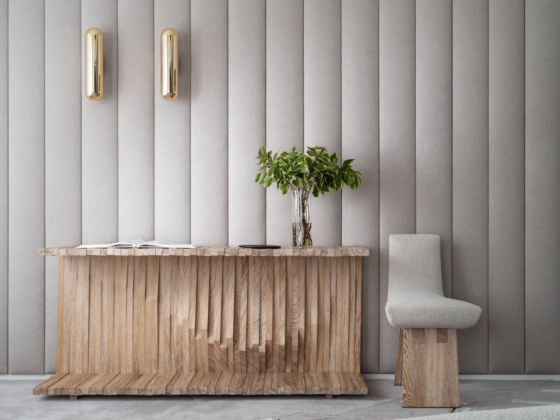 A modern entry console table made of wooden slats with a vase of green plants and a book on top, next to a textured white cha