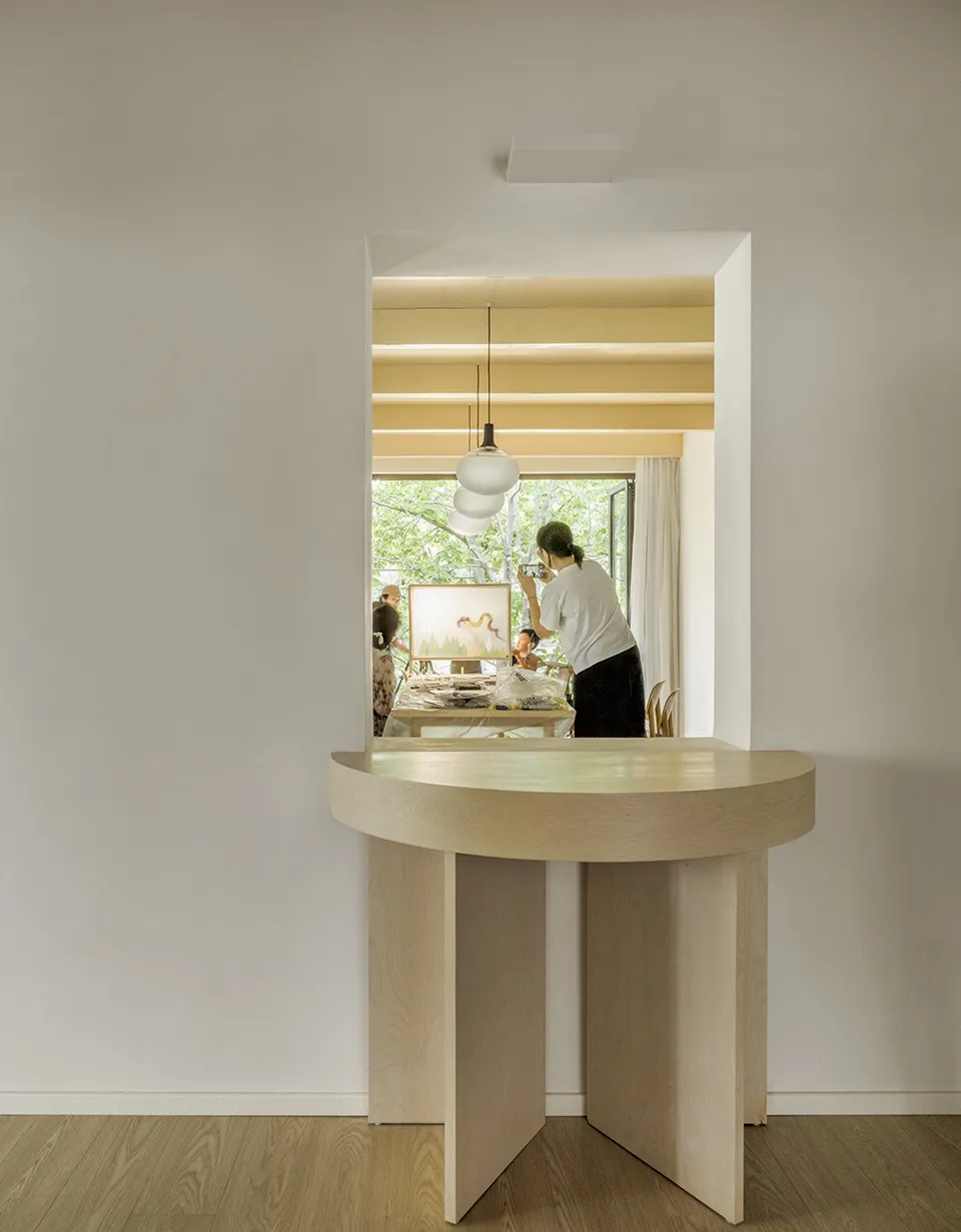 A minimalist home office featuring a wooden desk with an open storage in the center, a chair, and a view of a room with a pai