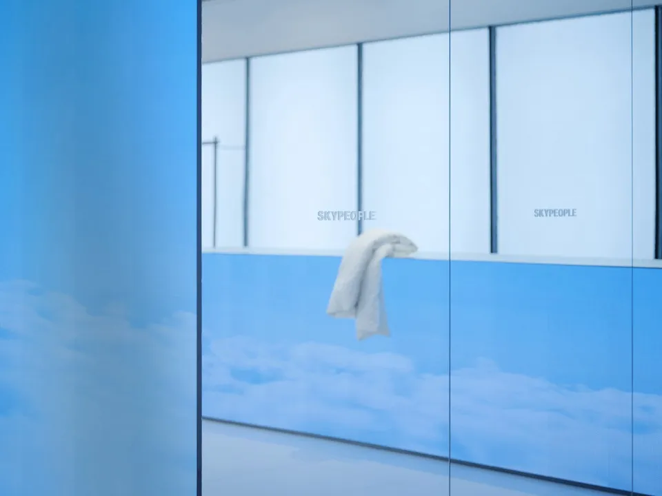A minimalist office space featuring transparent glass walls and a blue sky and clouds background.
