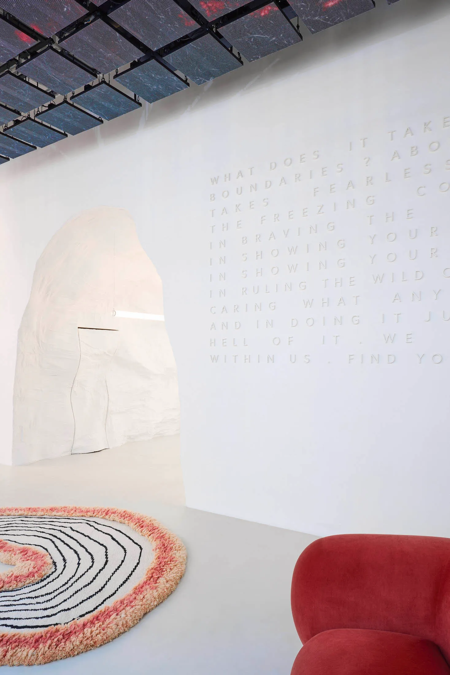 A minimalist white room with a textured white structure, a circular rug with black and pink lines, and a red upholstered chai