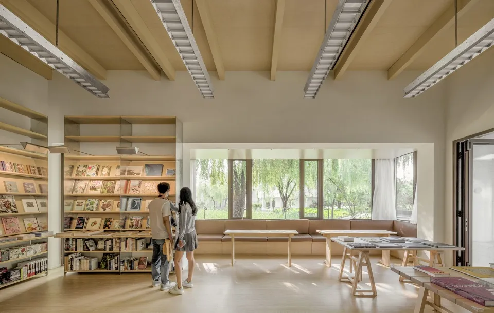People standing in a modern library interior with wooden shelves and tables, large windows, and a view of greenery outside.