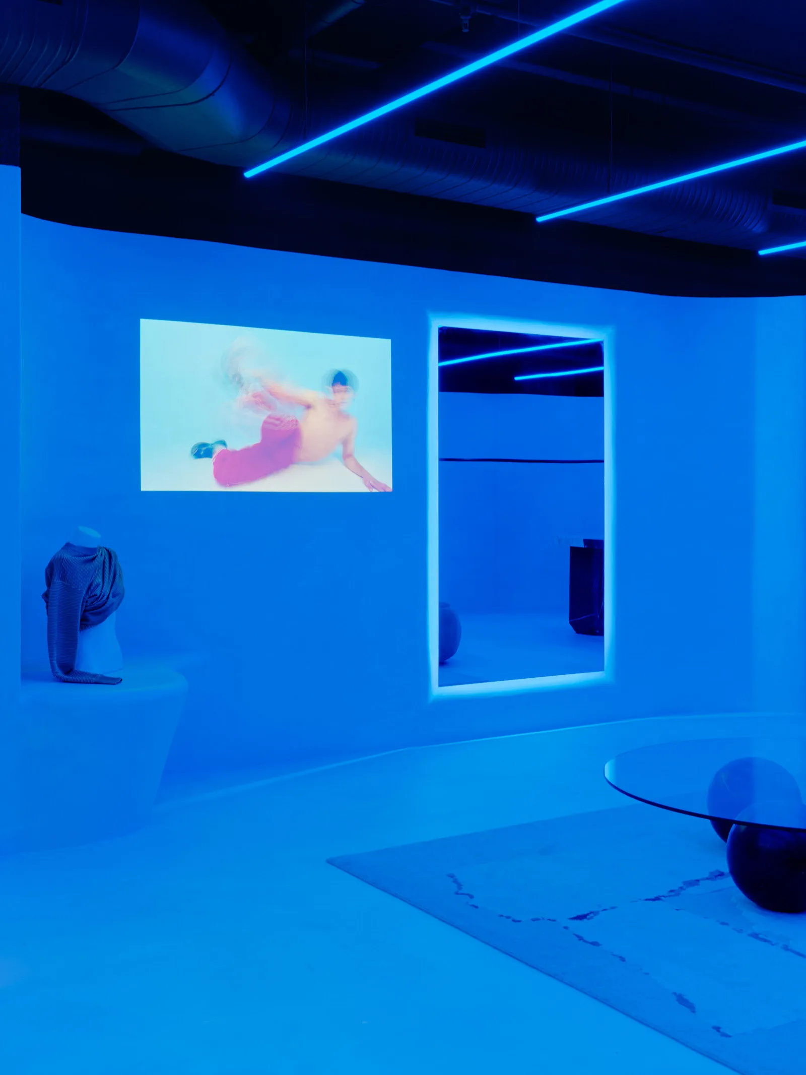 A minimalist installation with blue lighting, a mirror, and a projected image of a person on a screen.