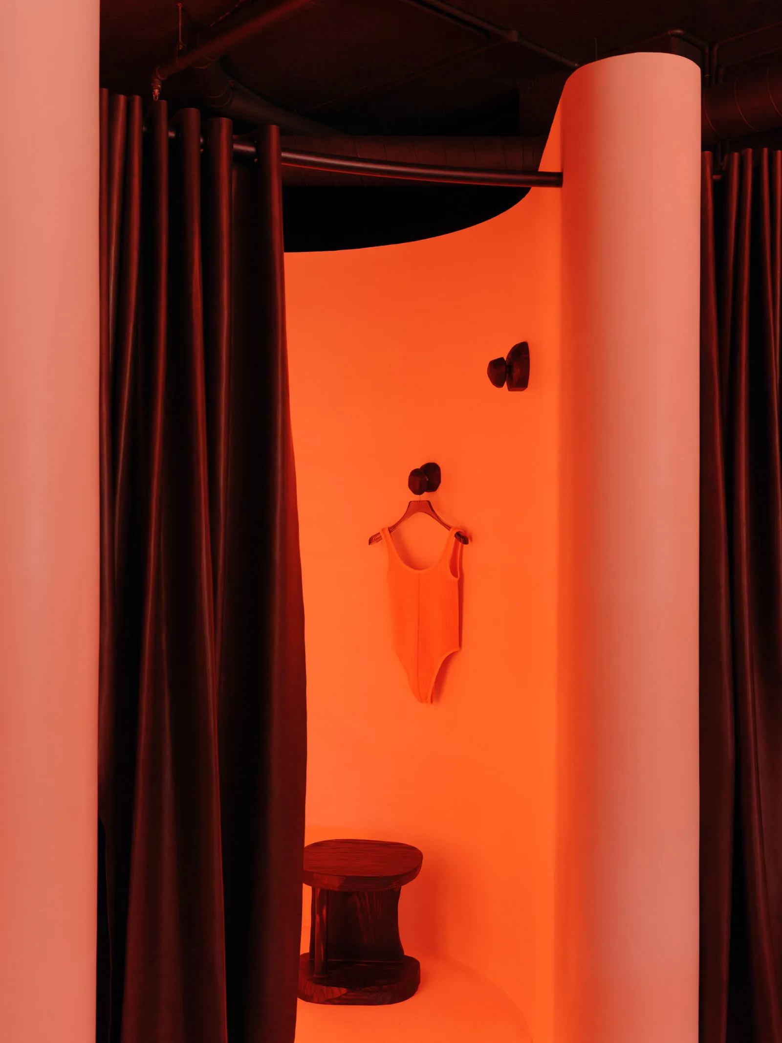 Clothing shop dressing room with bright orange lighting, dark curtains, white walls, and a round wooden stool inside.