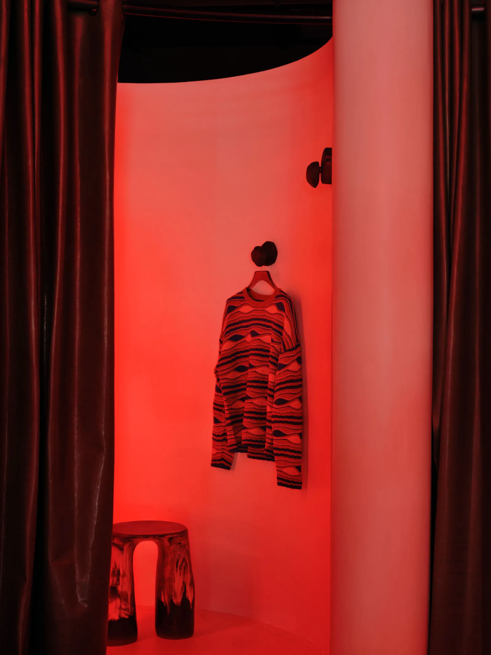 A red-lit interior space displaying clothing on a hanger, with a stool in the foreground.