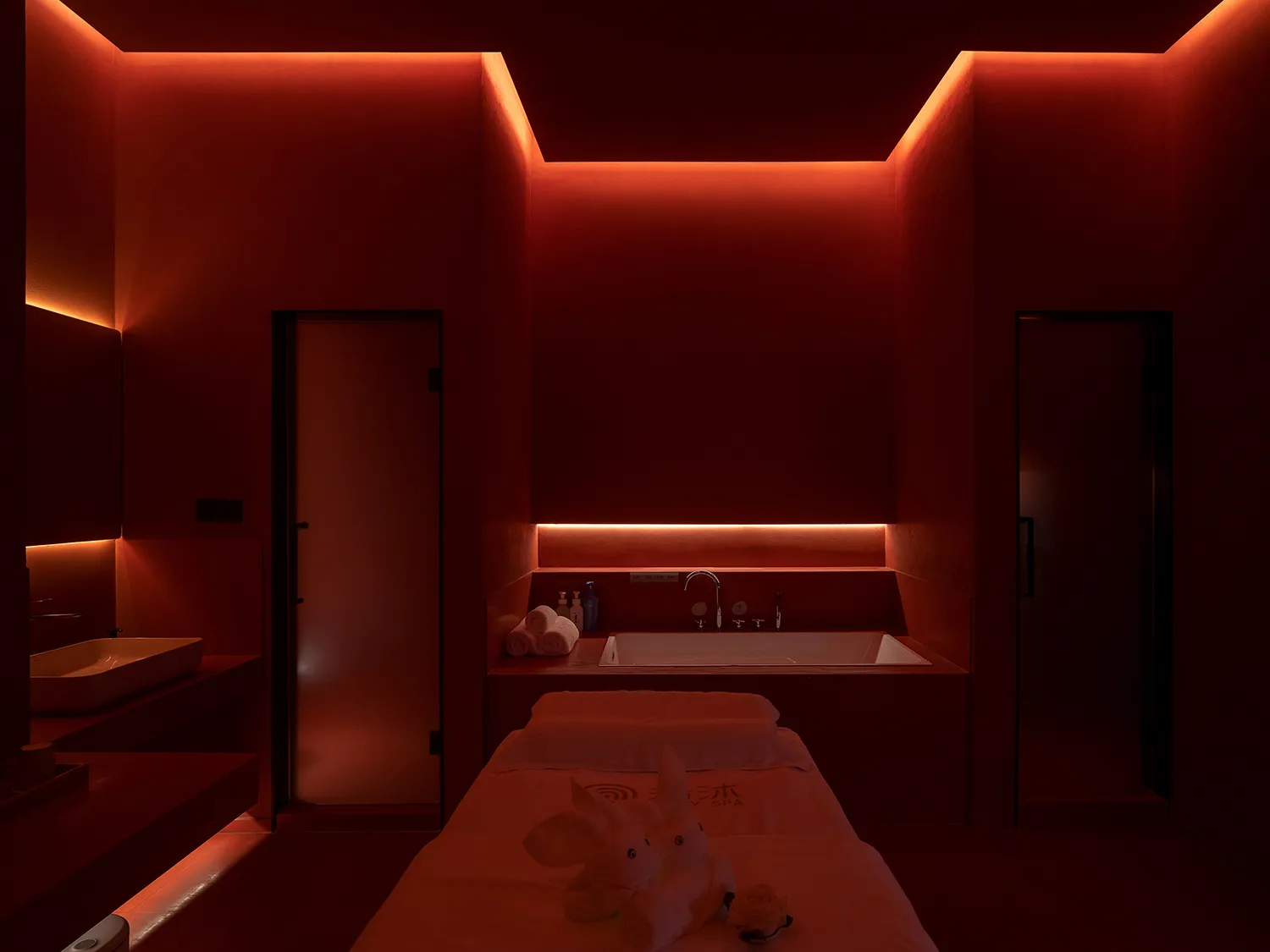 A spa room featuring a massage table with plush towels and a bathtub under dim red lighting.