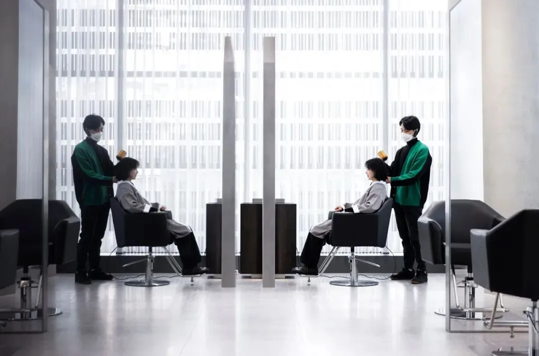 Modern salon setting with clients seated in black chairs, stylists cutting hair.