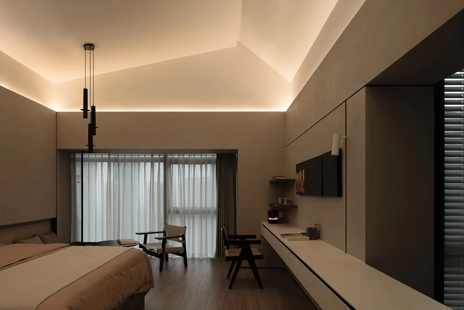 A modern minimalist bedroom with neutral-toned materials, featuring a bed, a desk, chairs, and a wall-mounted work area.