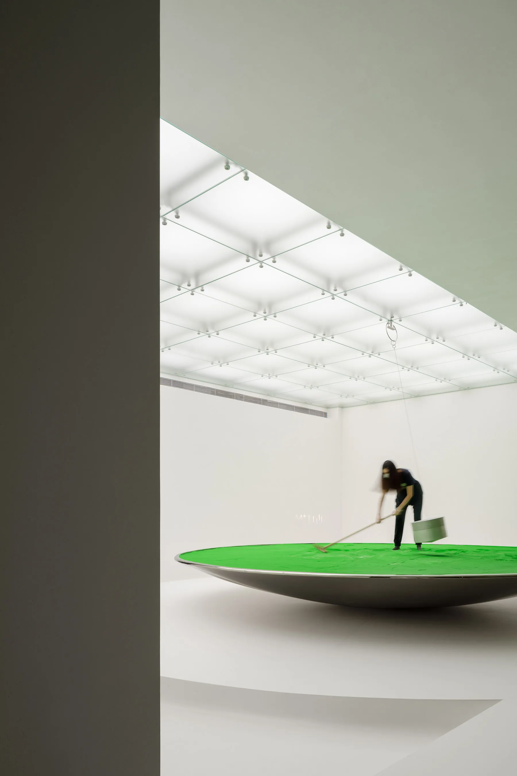 A minimalist art installation featuring a green surface being raked within a metallic bowl.