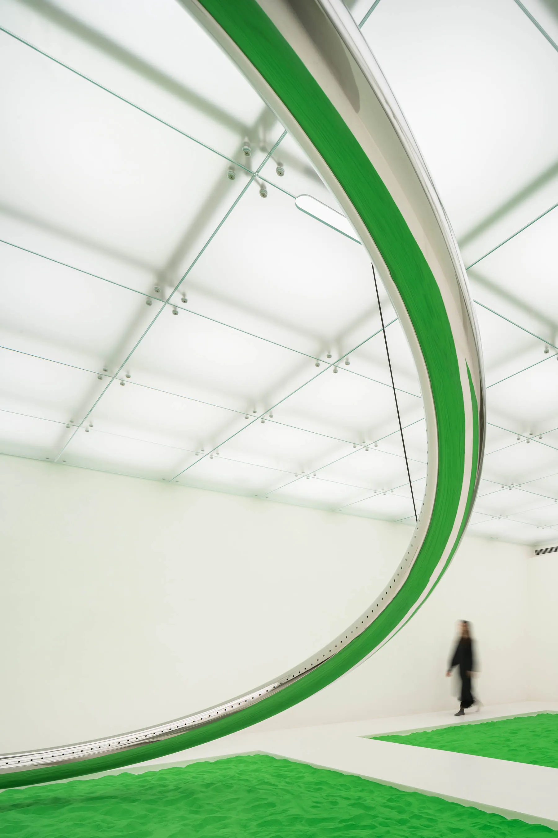 A modern art installation featuring a large, curved, metallic structure with organic green material suspended from the ceilin