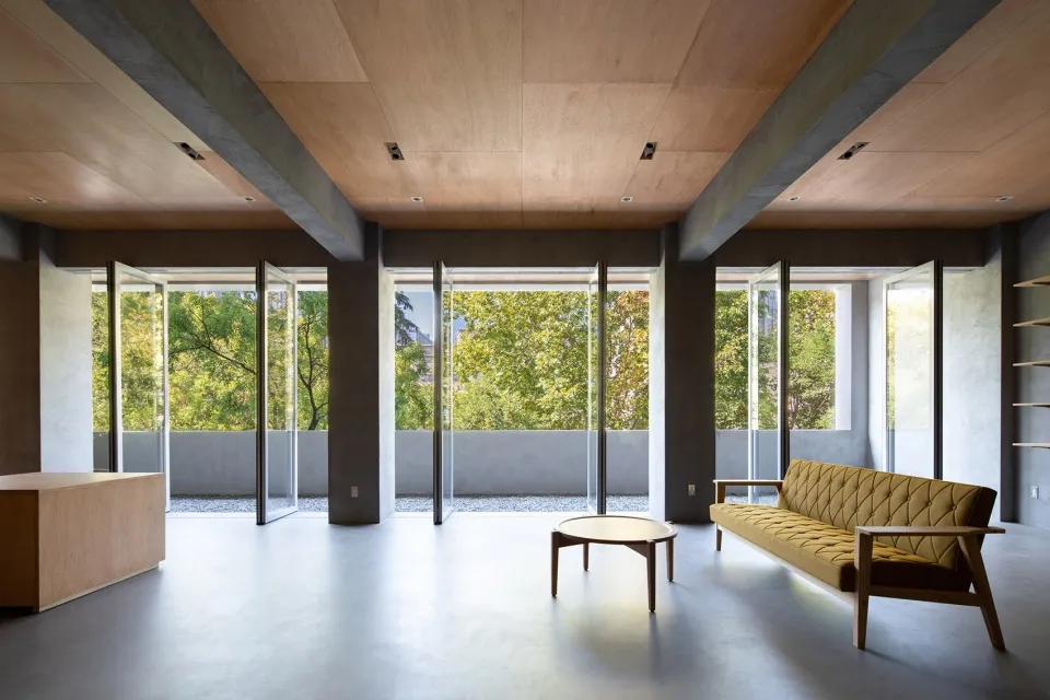 Modern living room with concrete and wooden elements, featuring large windows and a comfortable seating area.