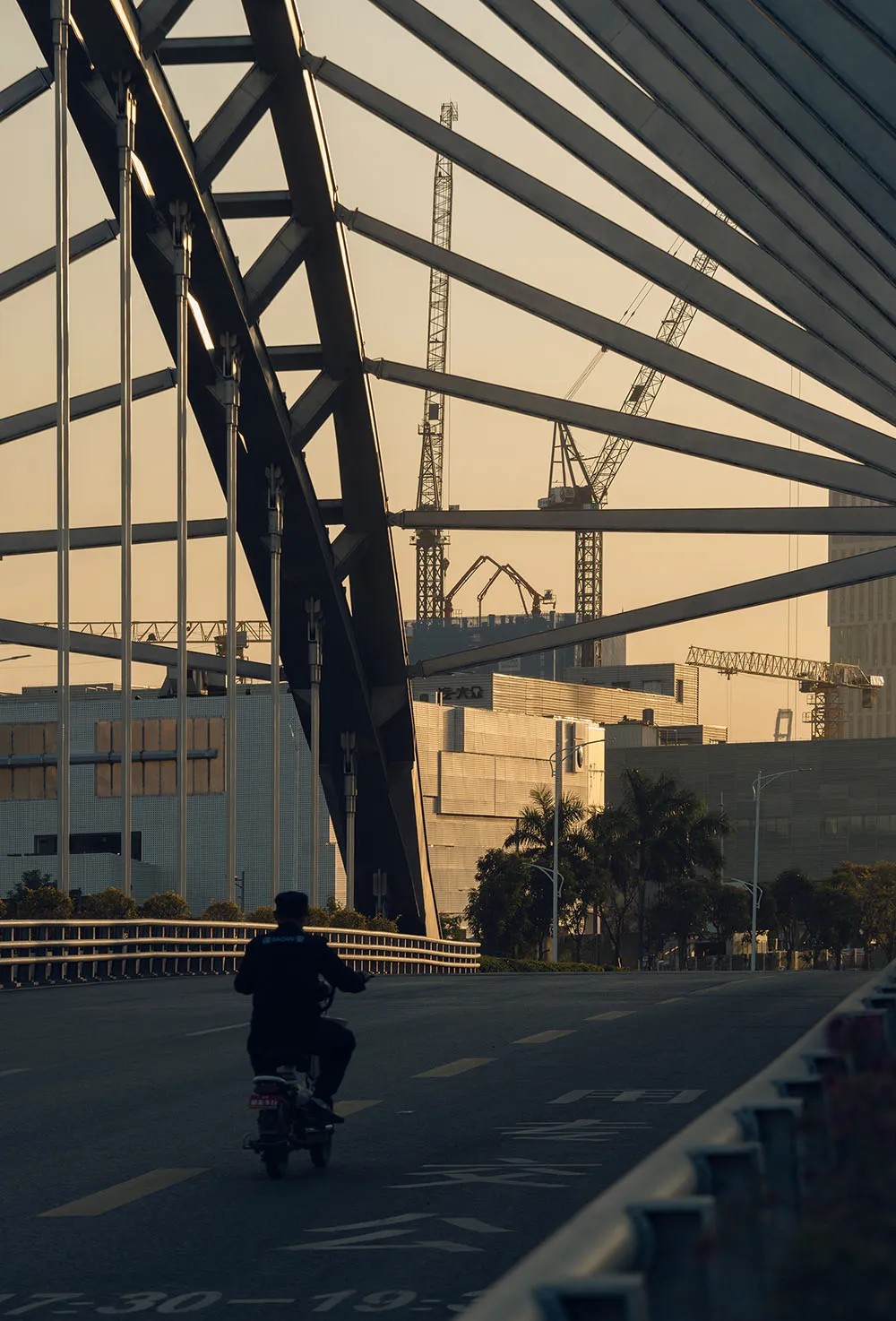 Person riding motorcycle under steel trusses during sunset at a construction site.