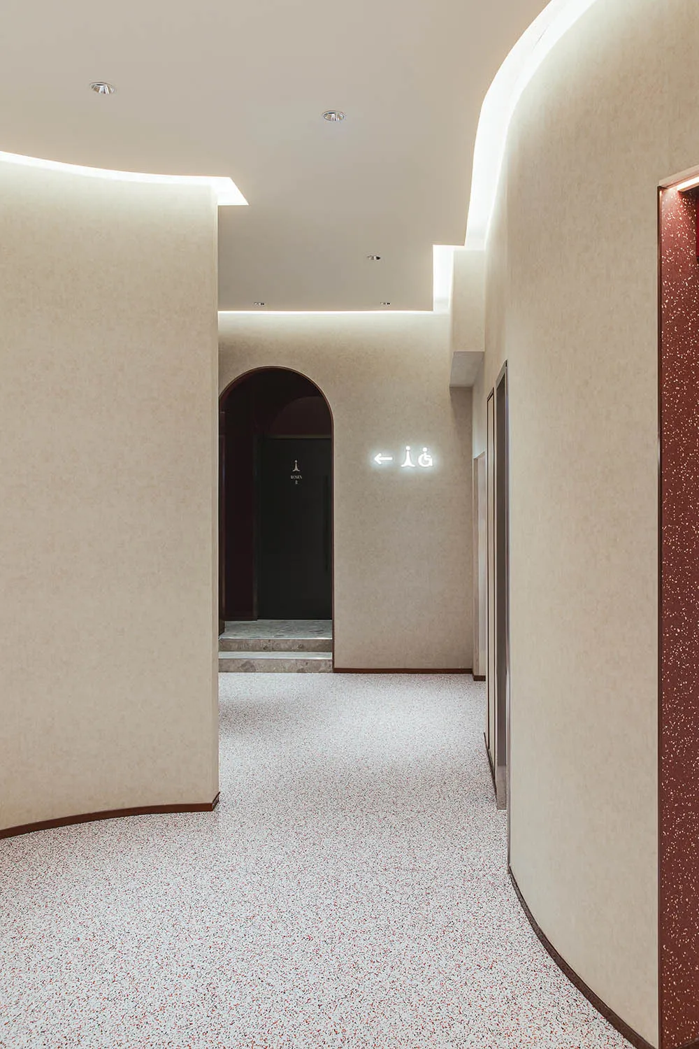 A minimalistic modern corridor with textured white walls and floor, featuring an arched doorway and functional signage.